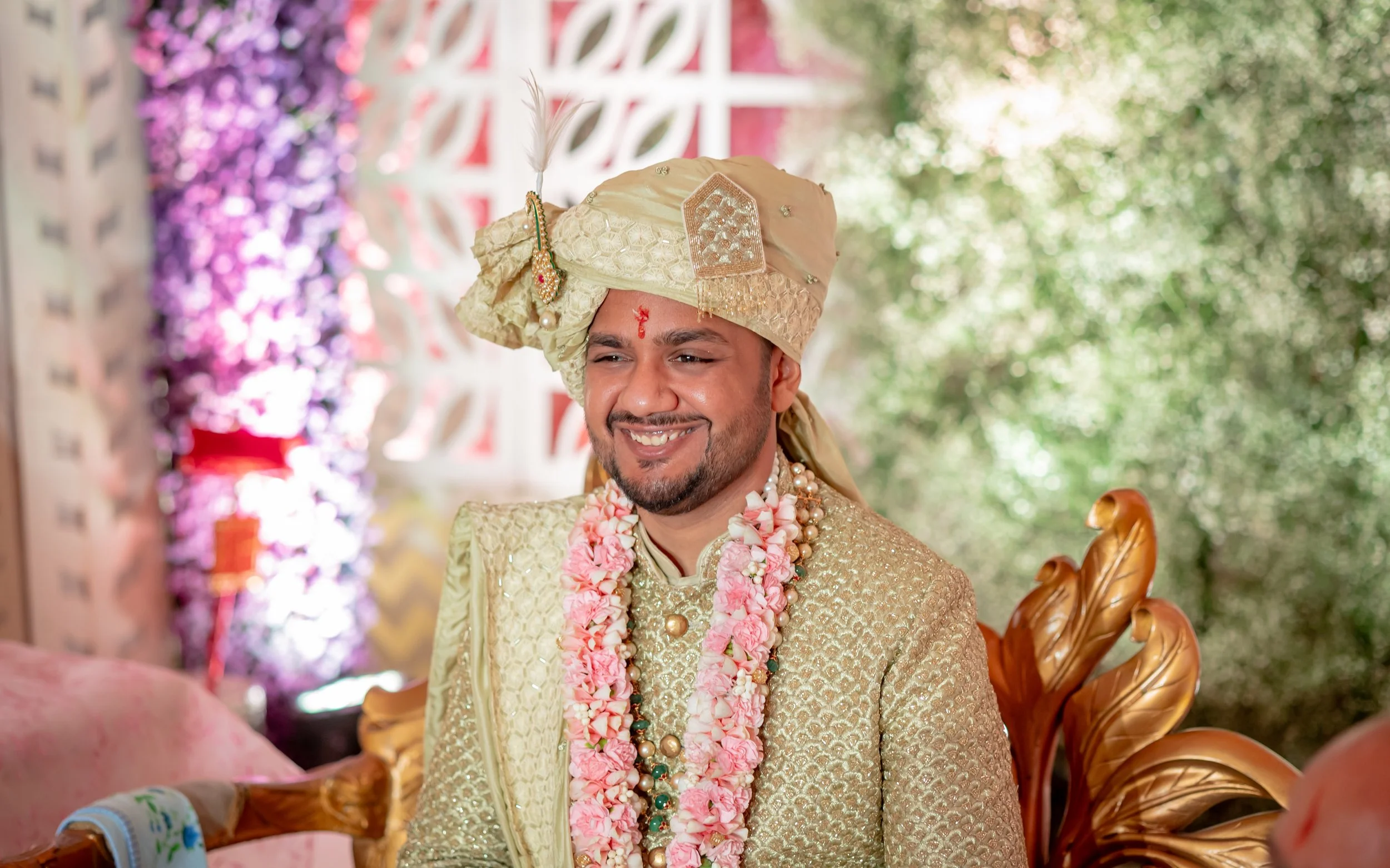 A groom dressed in traditional Indian wedding attire, wearing a gold sherwani and matching turban, adorned with multiple flower garlands and jewelry, smiling during his wedding ceremony.