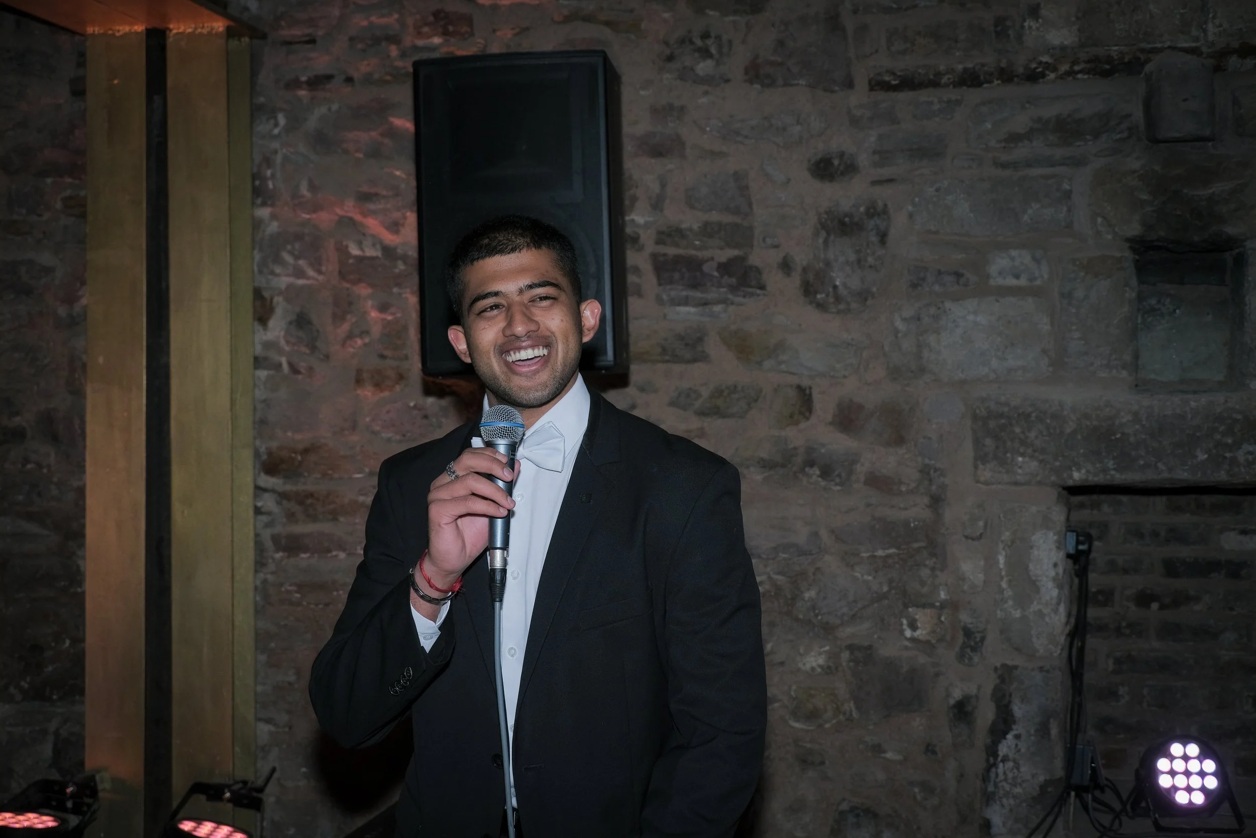 A young man in a dark suit and white shirt holding a microphone while smiling, standing in front of a stone wall with speakers and lighting equipment.