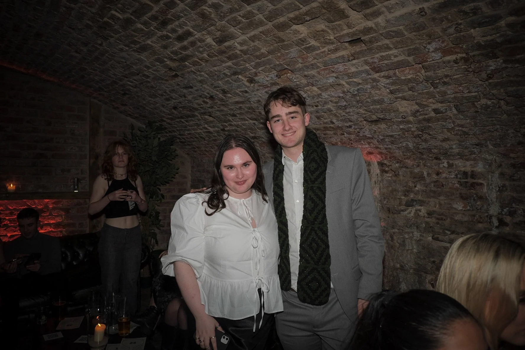 A young woman wearing a white blouse and a young man in a gray suit with a patterned scarf standing together in a brick-walled, dimly lit indoor setting, possibly a bar or restaurant, with other people in the background.
