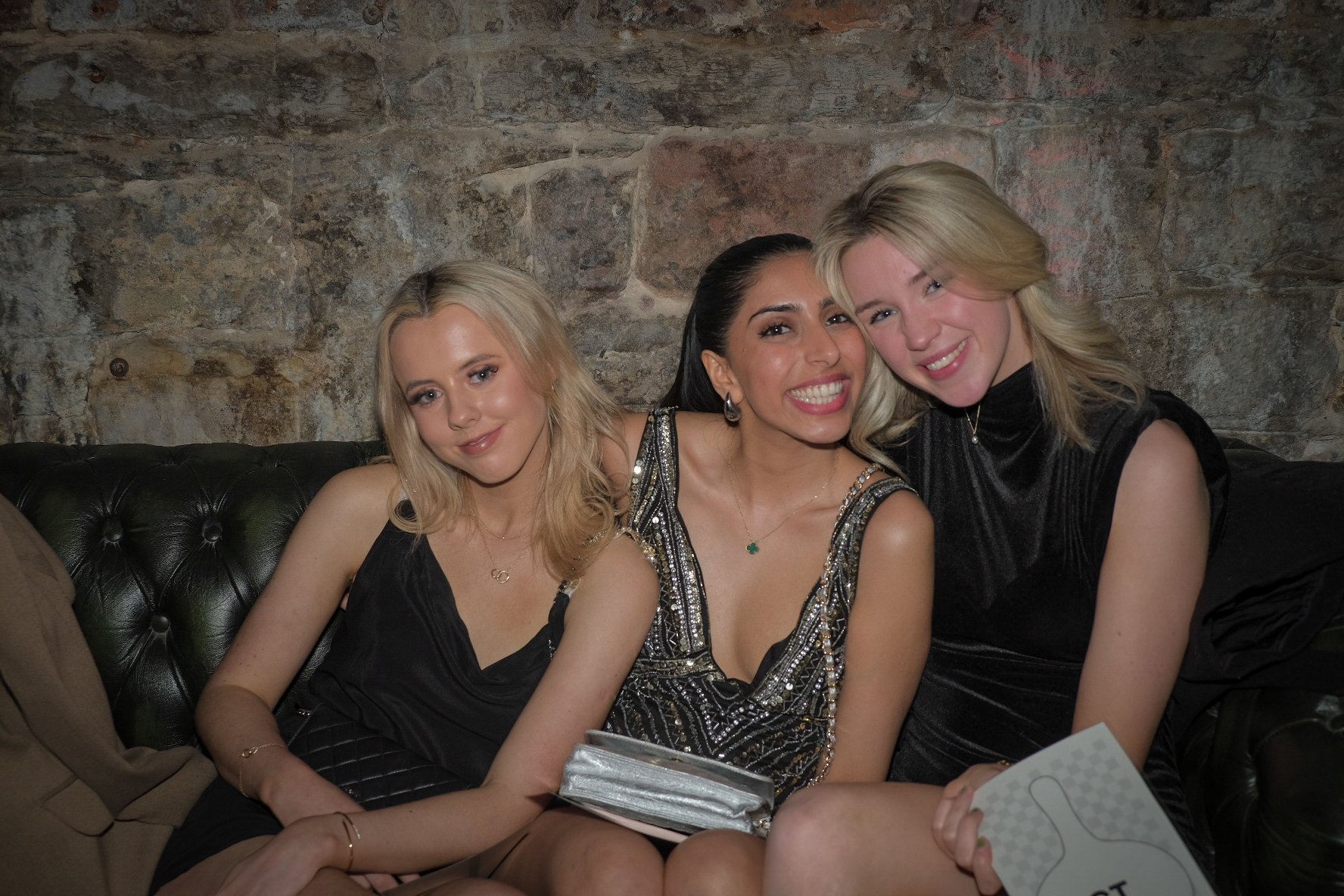 Three women sitting on a black leather couch, smiling and posing for a photo against a stone wall background. They are dressed in black evening attire.