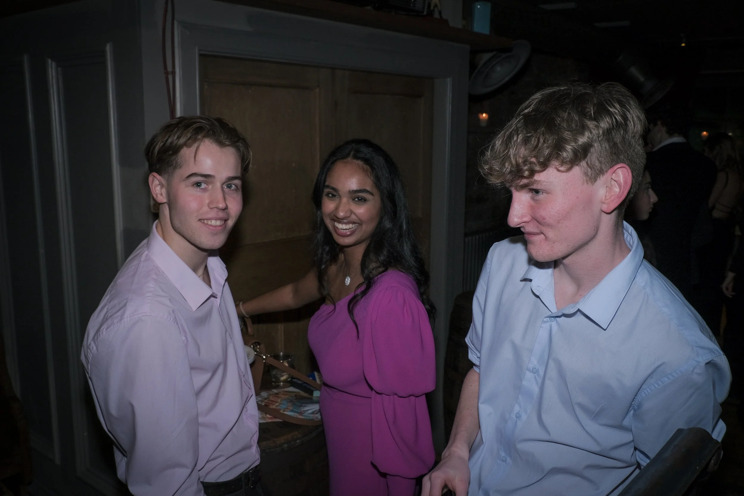 Three young people, two men and one woman, standing indoors at a social event. The woman wears a pink dress, the man on the left has a light pink shirt, and the man on the right has a light blue shirt. They are smiling and facing the camera.