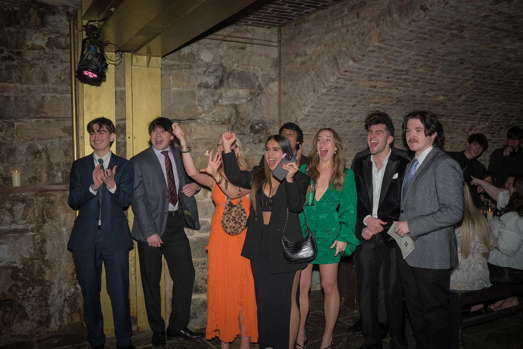 Group of people dressed in formal attire enjoying a celebration or party in a dimly lit venue with stone walls.