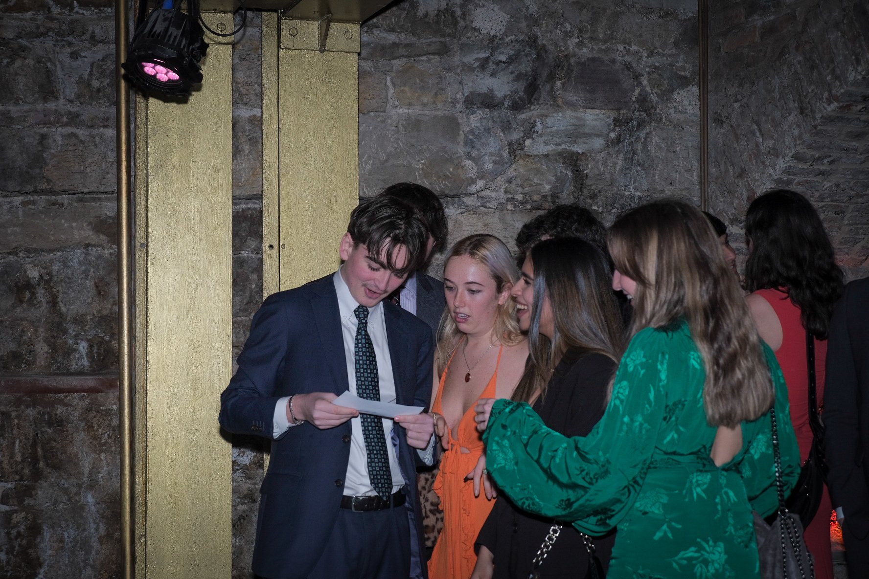 Group of young people dressed in formal wear at a party or event, gathered around and looking at a piece of paper, in a dimly lit room with stone walls.