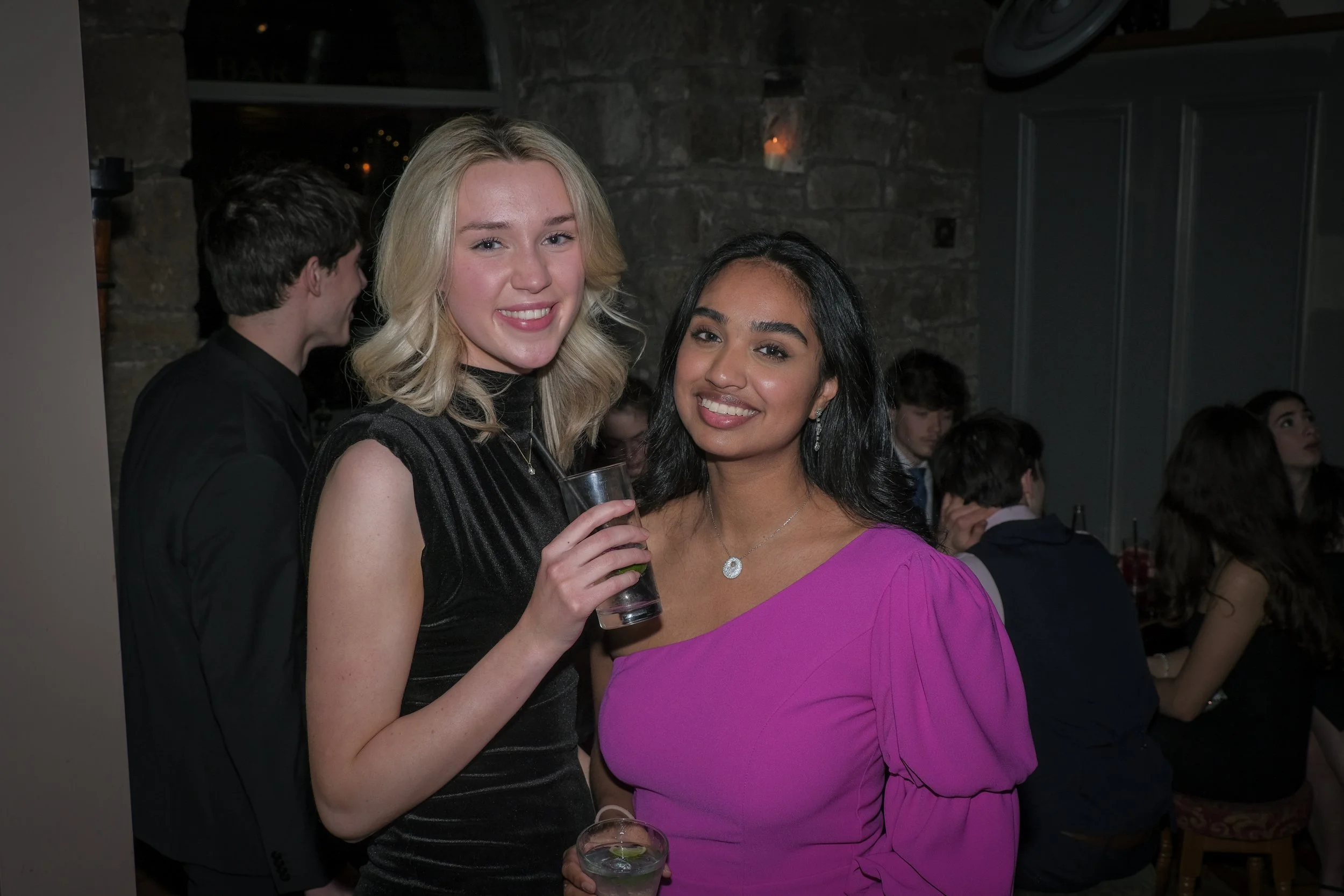 Two women smiling at a party, one holding a drink and wearing a black dress, the other wearing a pink dress and holding a clear drink.