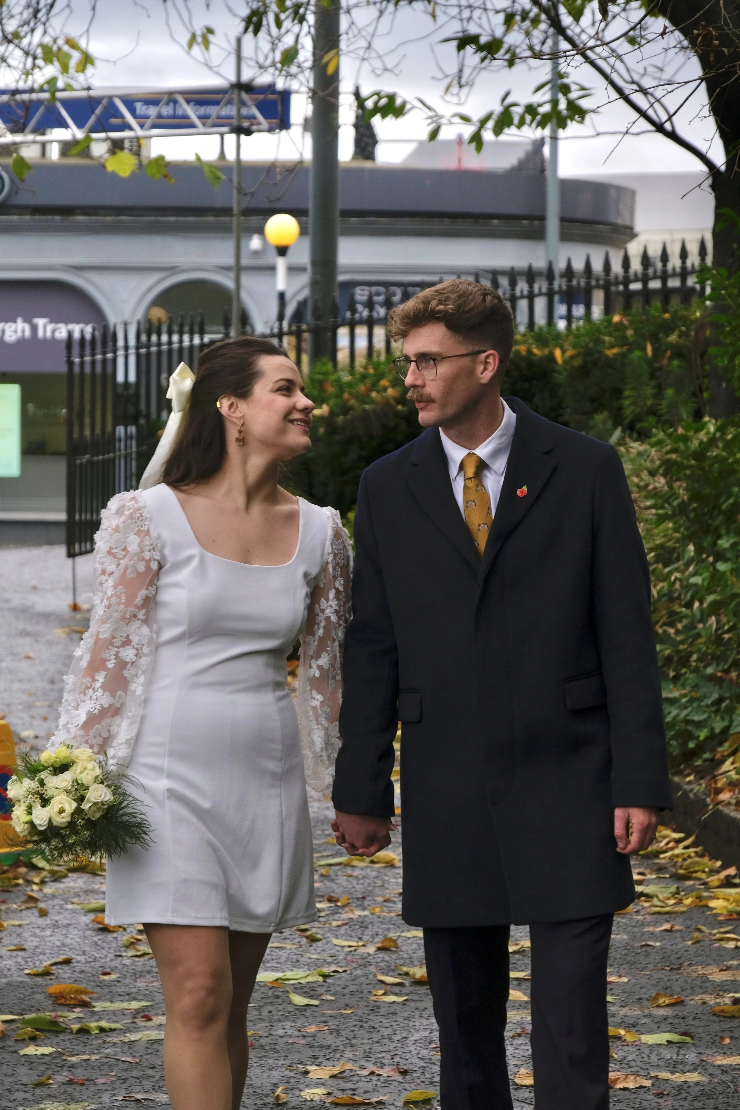 A couple, dressed in wedding attire, holding hands and walking outdoors on a leaf-covered path. The bride is smiling, wearing a white dress with lace sleeves, holding a bouquet, and has a white bow in her hair. The groom is in a dark coat with a must