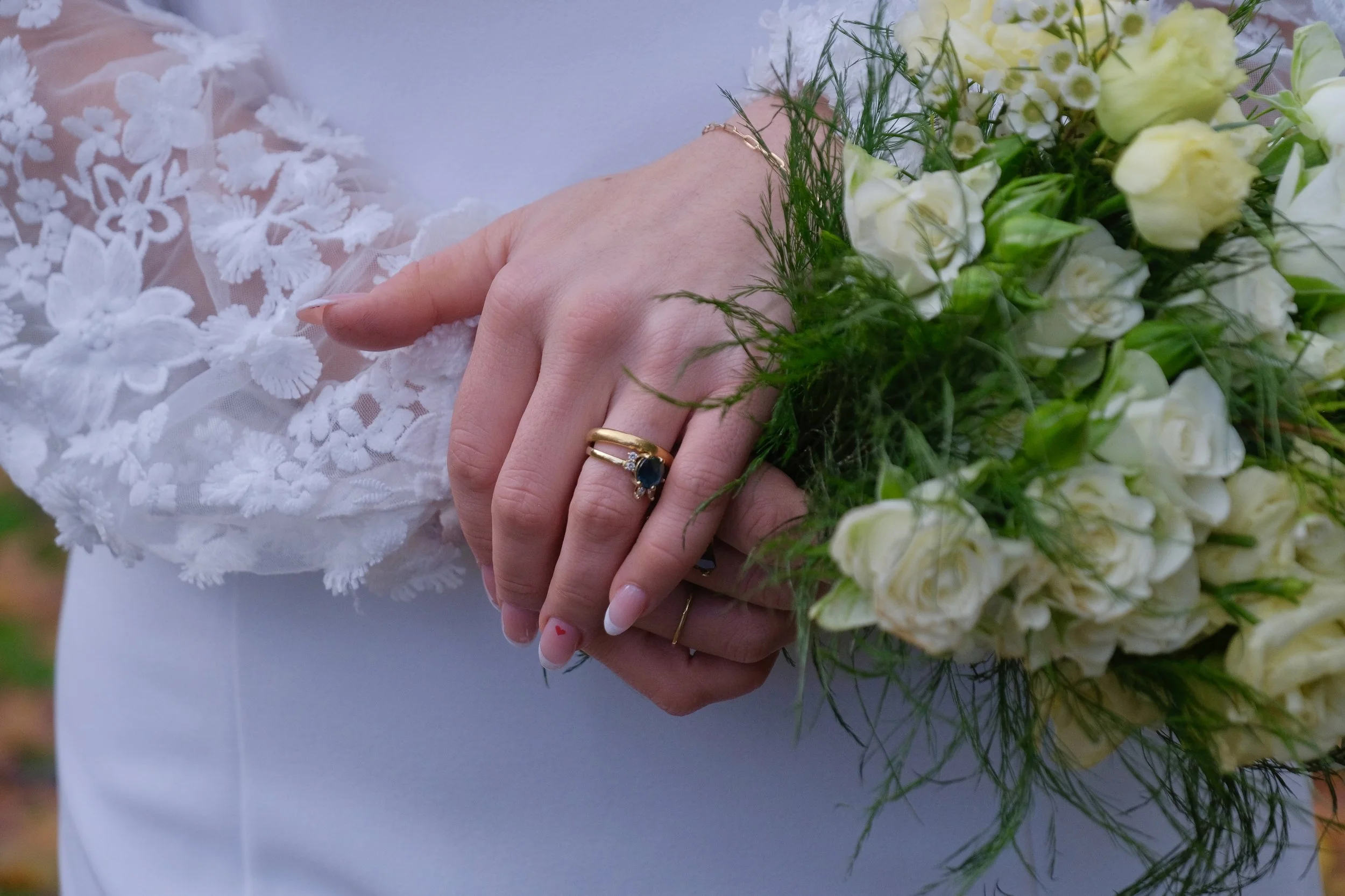 Close-up of a person holding a bouquet of white roses with green foliage, wearing a white dress with floral embroidery, a gold ring with a dark gemstone, and a delicate bracelet.