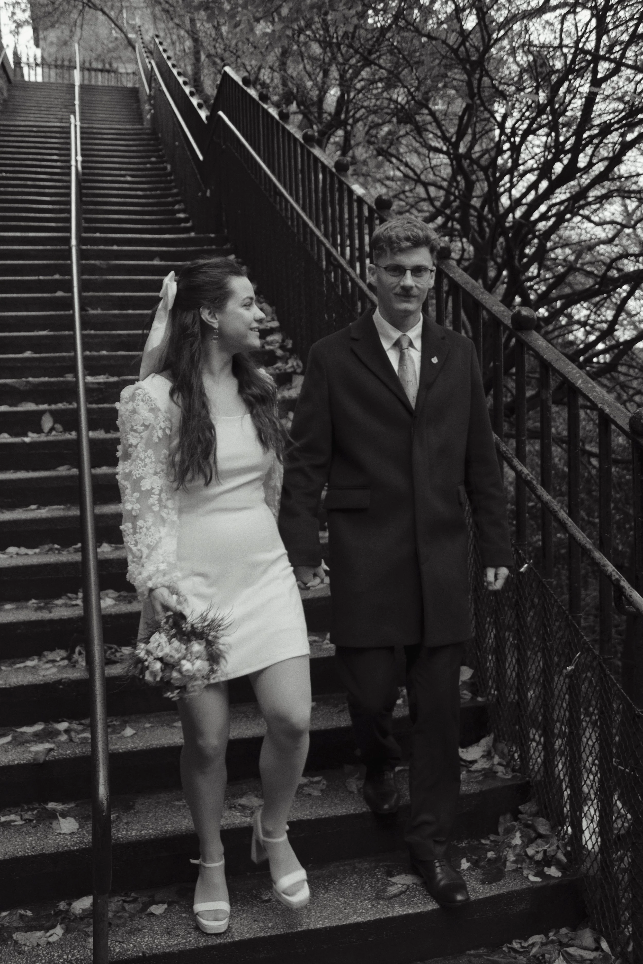 A black and white photo of a bride and groom walking down outdoor stairs holding hands, with trees and fallen leaves in the background.