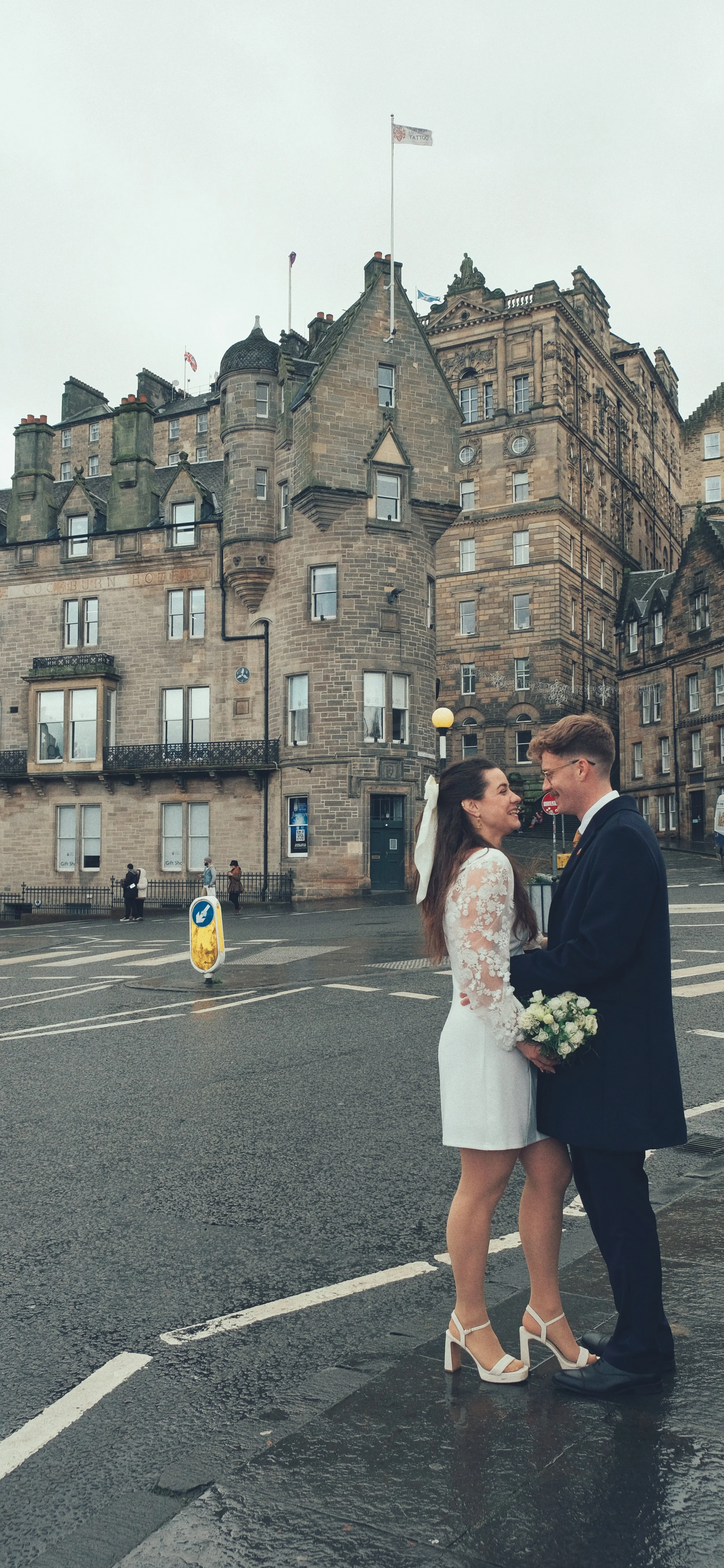 A newlywed couple stand face to face on a wet city street, smiling at each other, with the bride holding a bouquet of white roses. The bride wears a white lace dress and high heels, while the groom is dressed in a navy suit with a yellow tie. Behind 