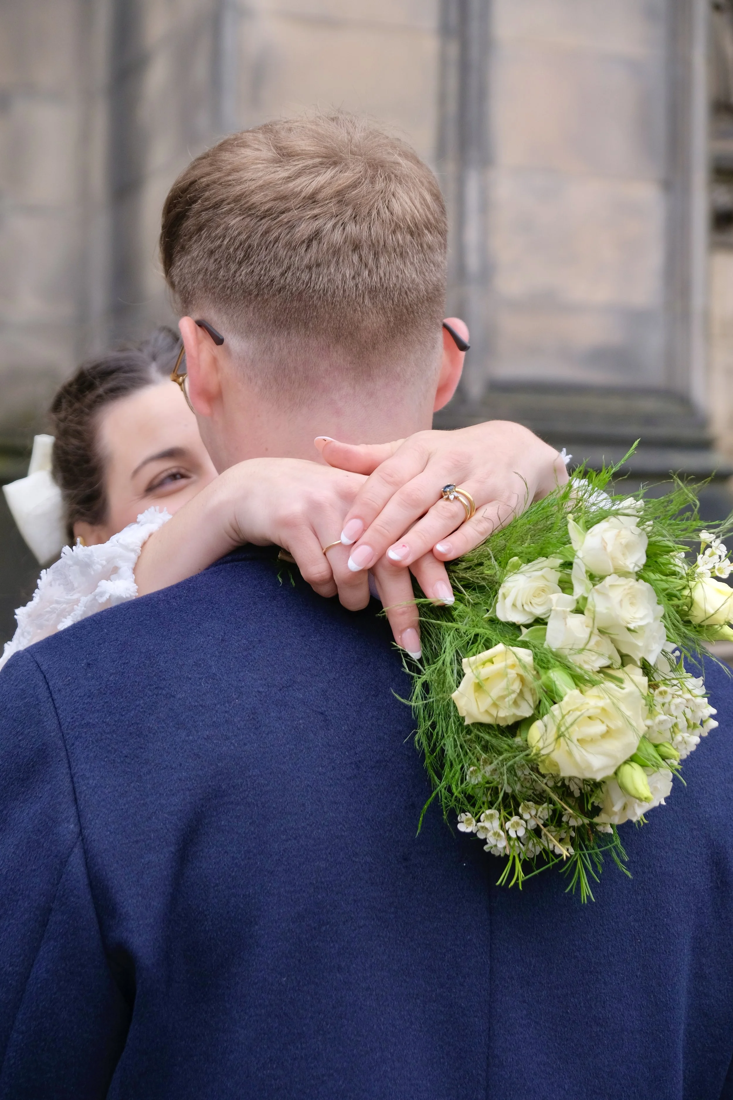 A bride hugging a groom from behind, holding a bouquet of white roses and greenery, during a wedding ceremony or celebration.