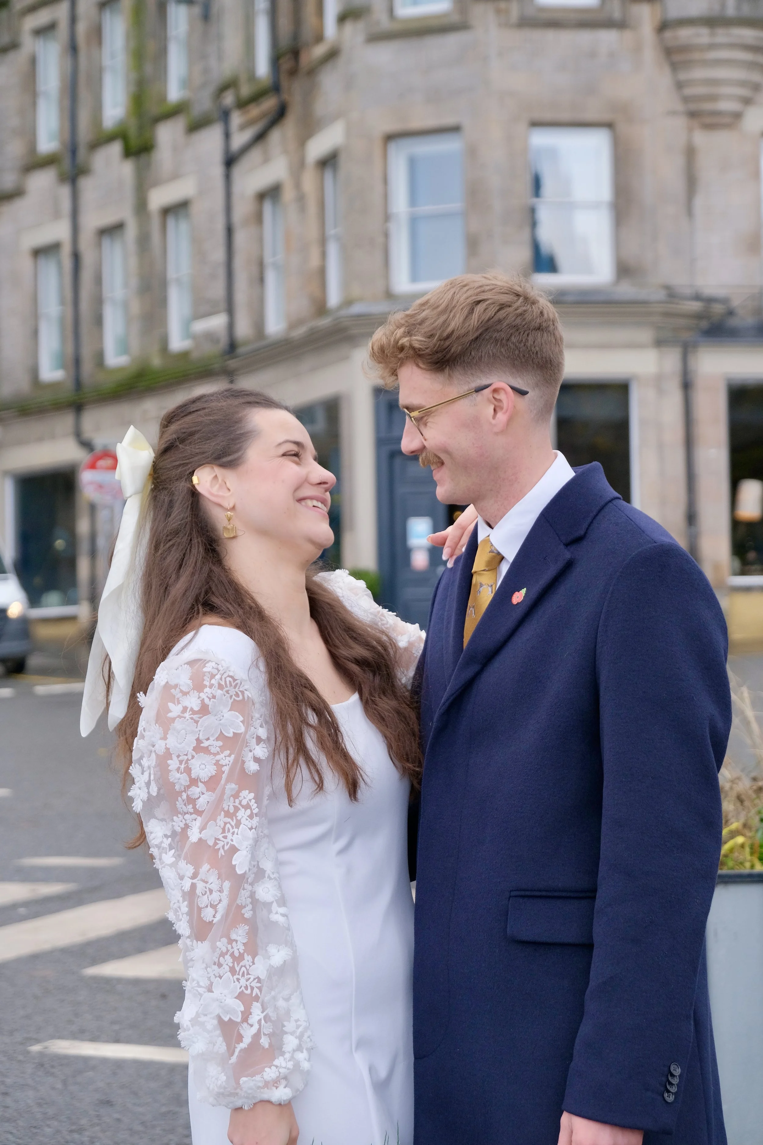 A smiling couple in wedding attire standing close together on a city street, looking at each other lovingly.