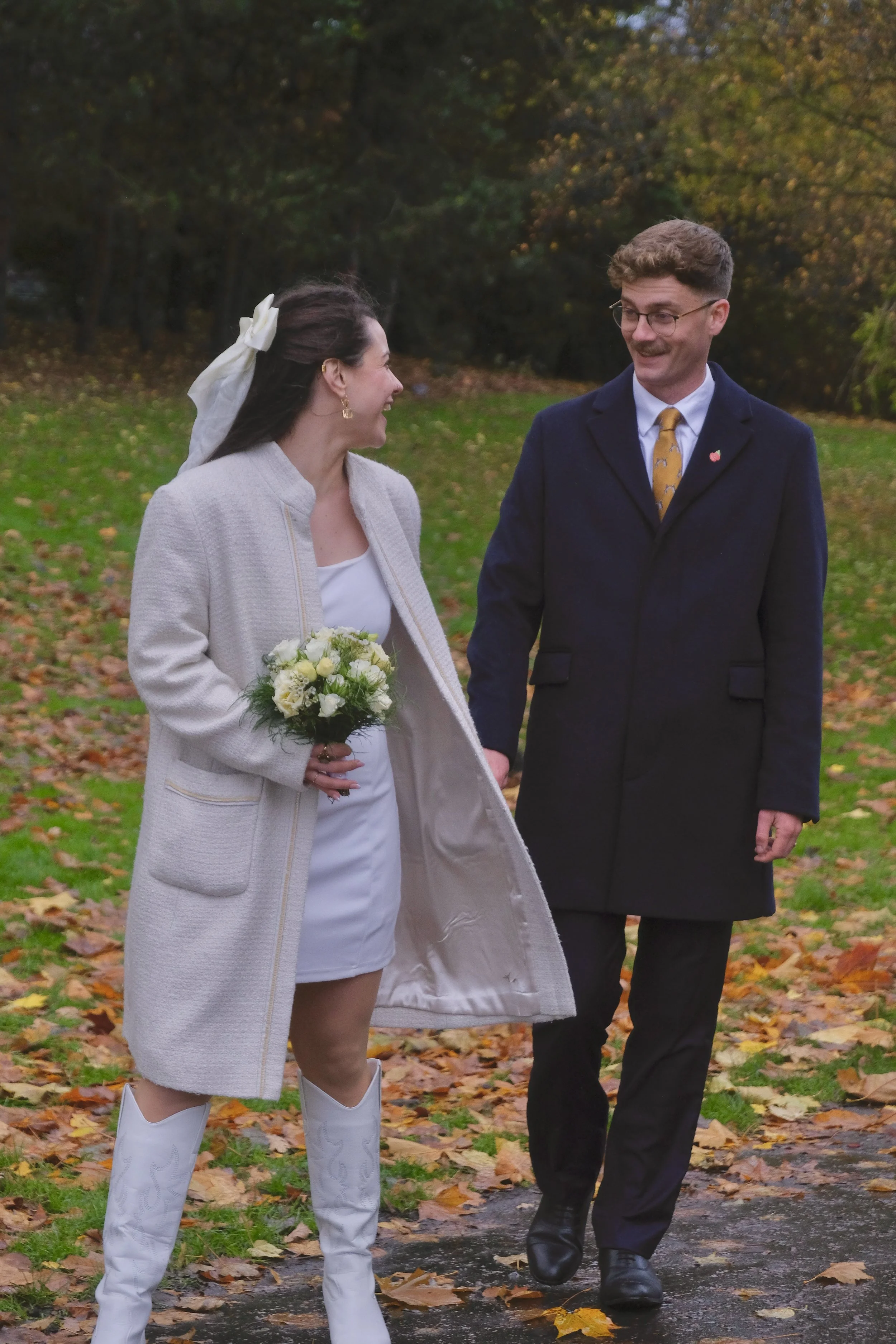 A woman in a white dress and white coat holding a bouquet of white flowers, walking outdoors with a man in a dark coat and yellow tie, in a park with fallen autumn leaves.