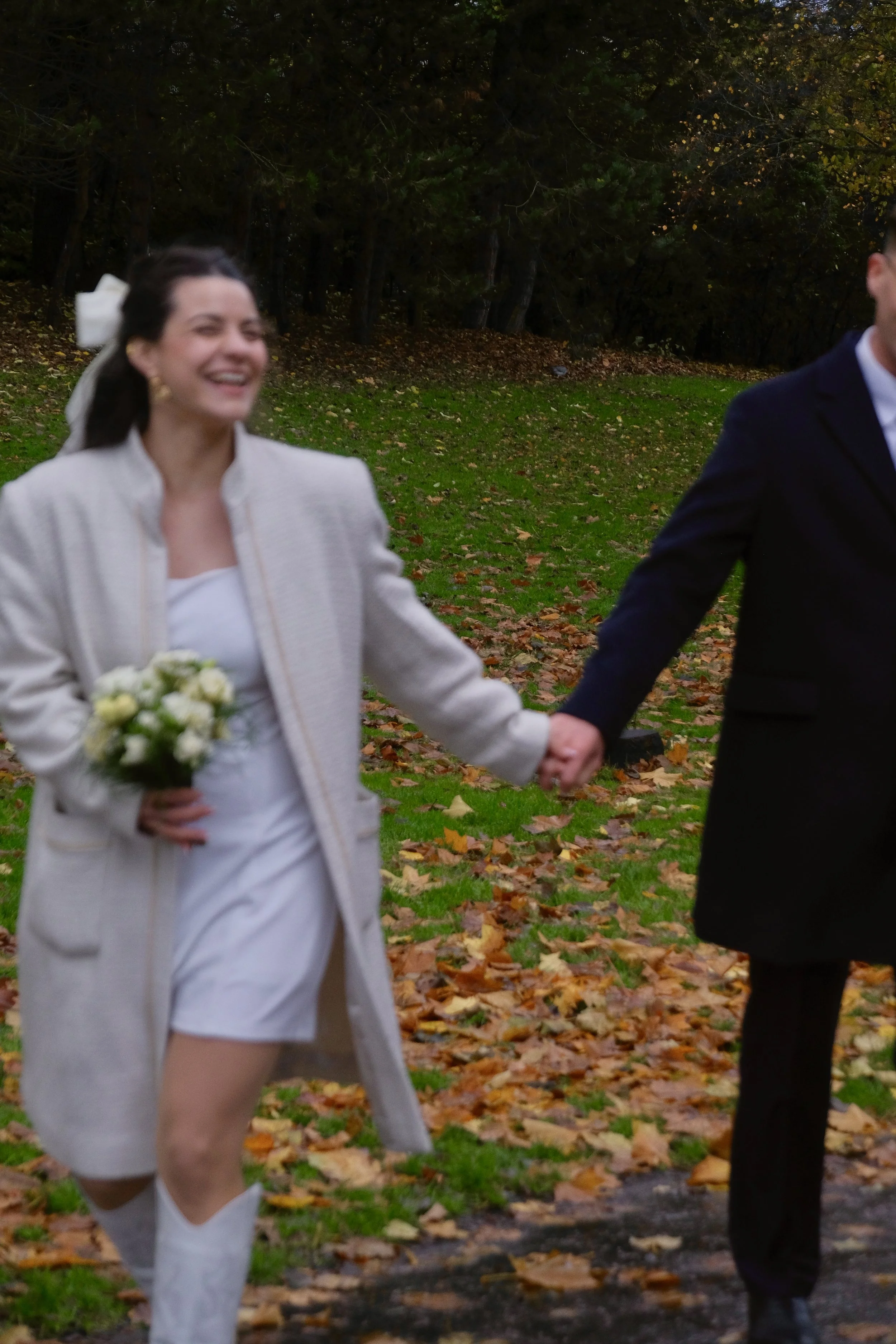A woman dressed in white holding a bouquet, smiling and holding hands with a man dressed in a dark suit, outdoors in a fall setting with fallen leaves on the ground.