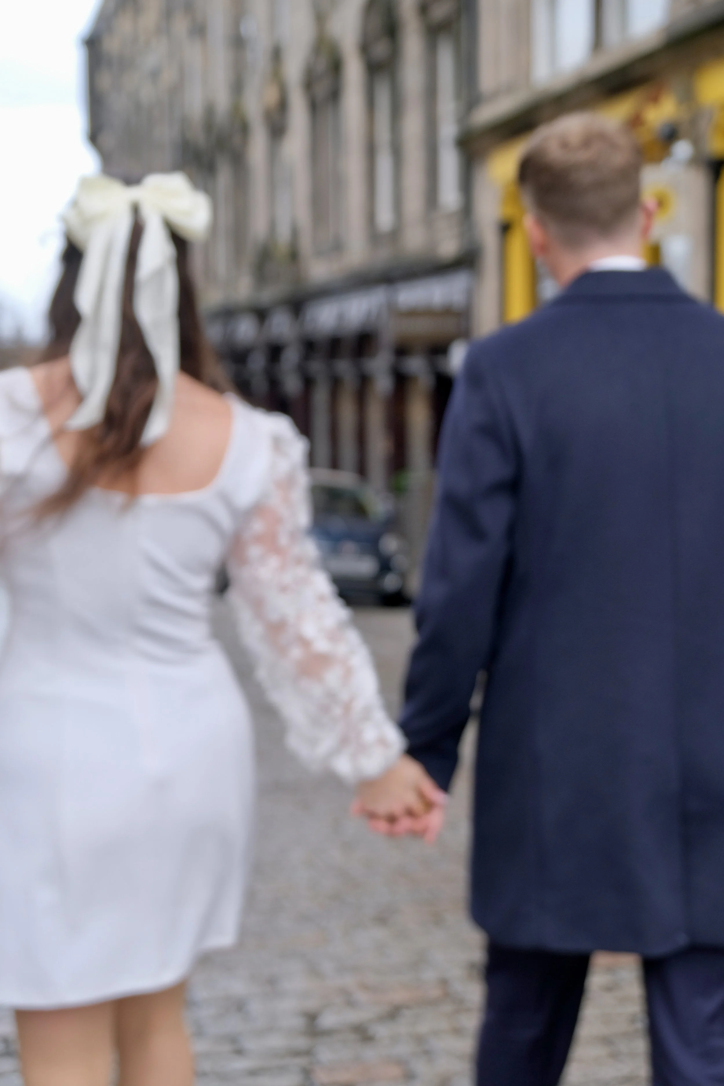 A couple walking hand in hand down a cobblestone street in a city, holding hands with a woman wearing a white dress and a large bow in her hair, and a man in a dark suit.
