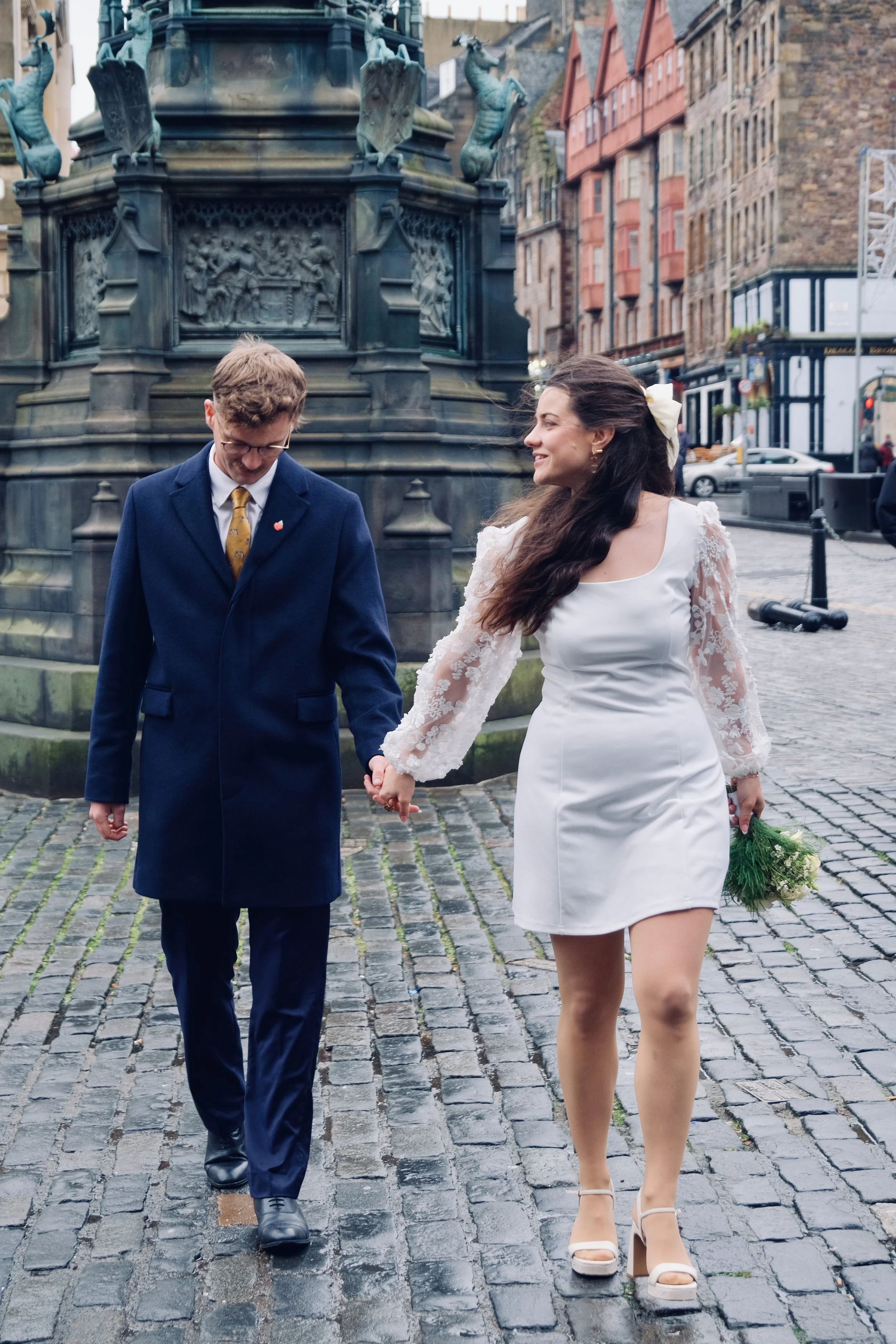 A couple dressed in wedding attire holding hands and walking on cobblestone street near a monument in an urban area.