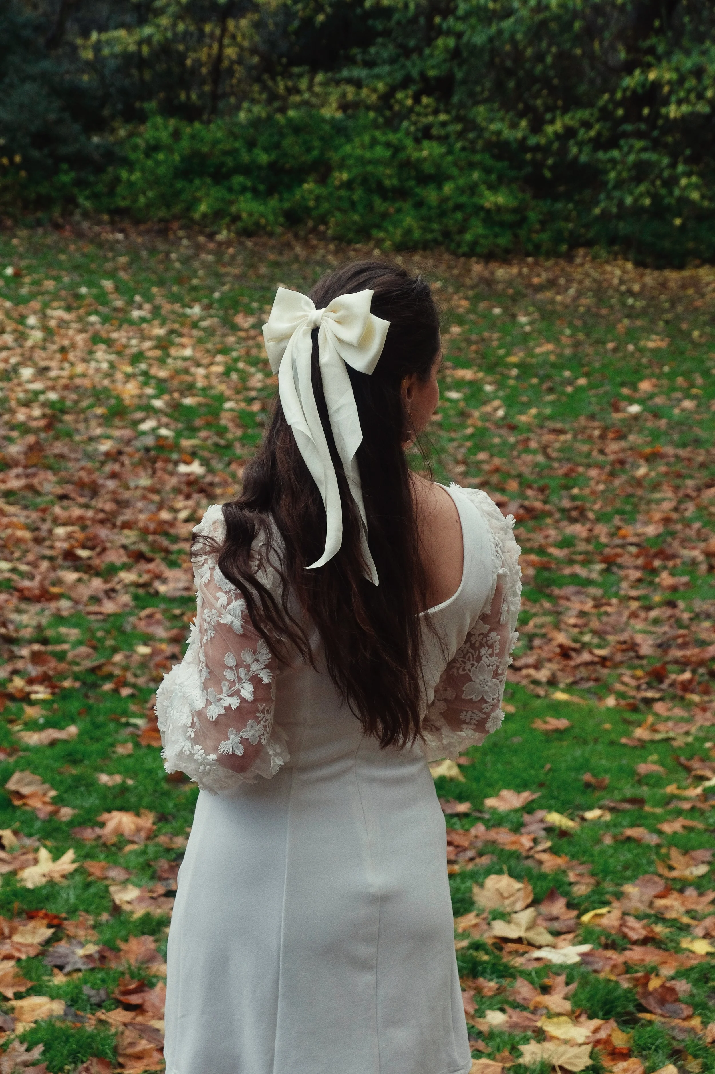 A woman with long dark hair wearing a white dress with lace sleeves, standing outdoors on a fall day with leaves scattered on the grass, and a large white bow in her hair.