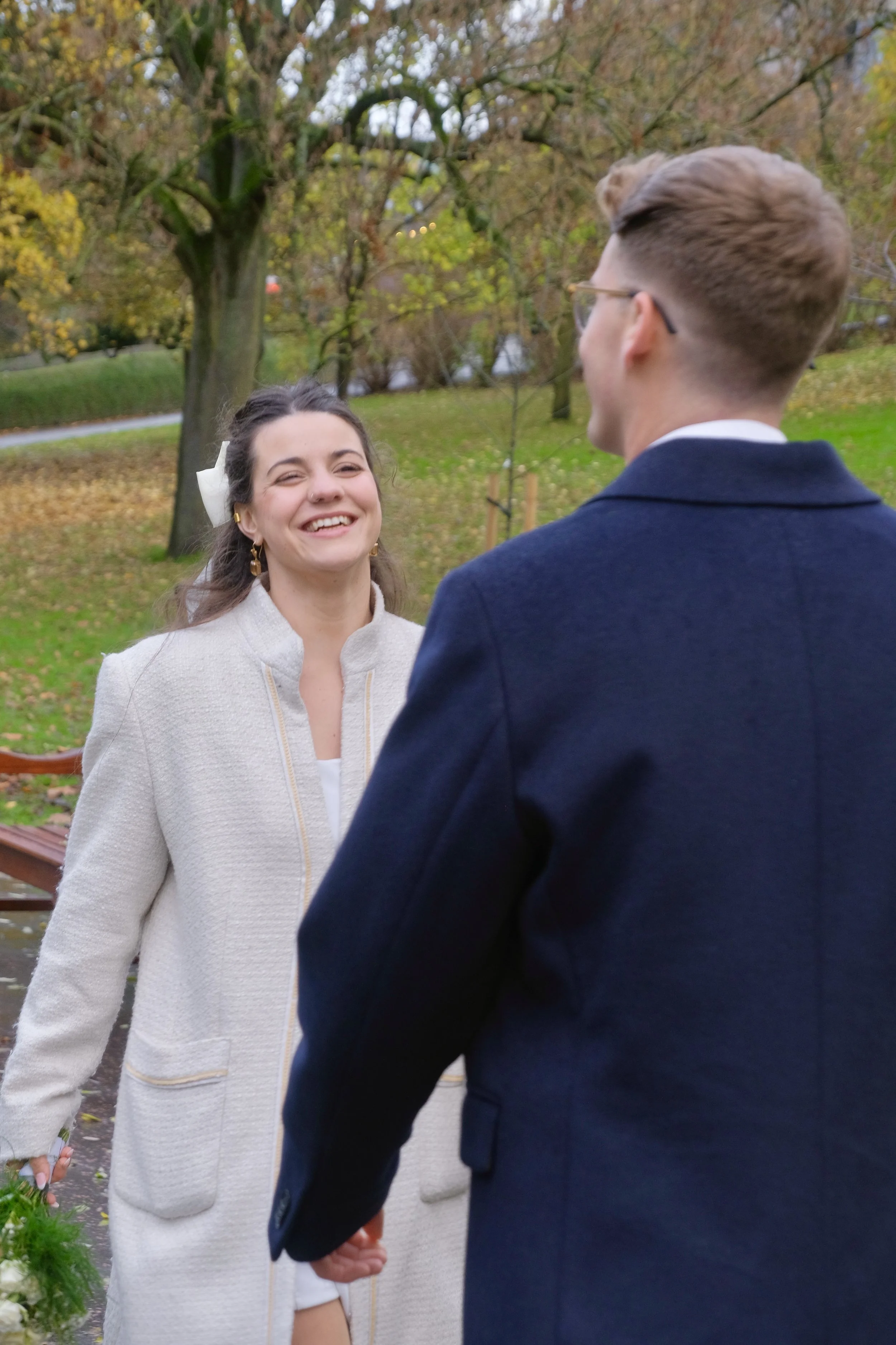 A woman smiling and holding hands with a man outdoors during fall. The woman is wearing a white coat and earrings, while the man is wearing a dark coat. There are trees with autumn leaves in the background.