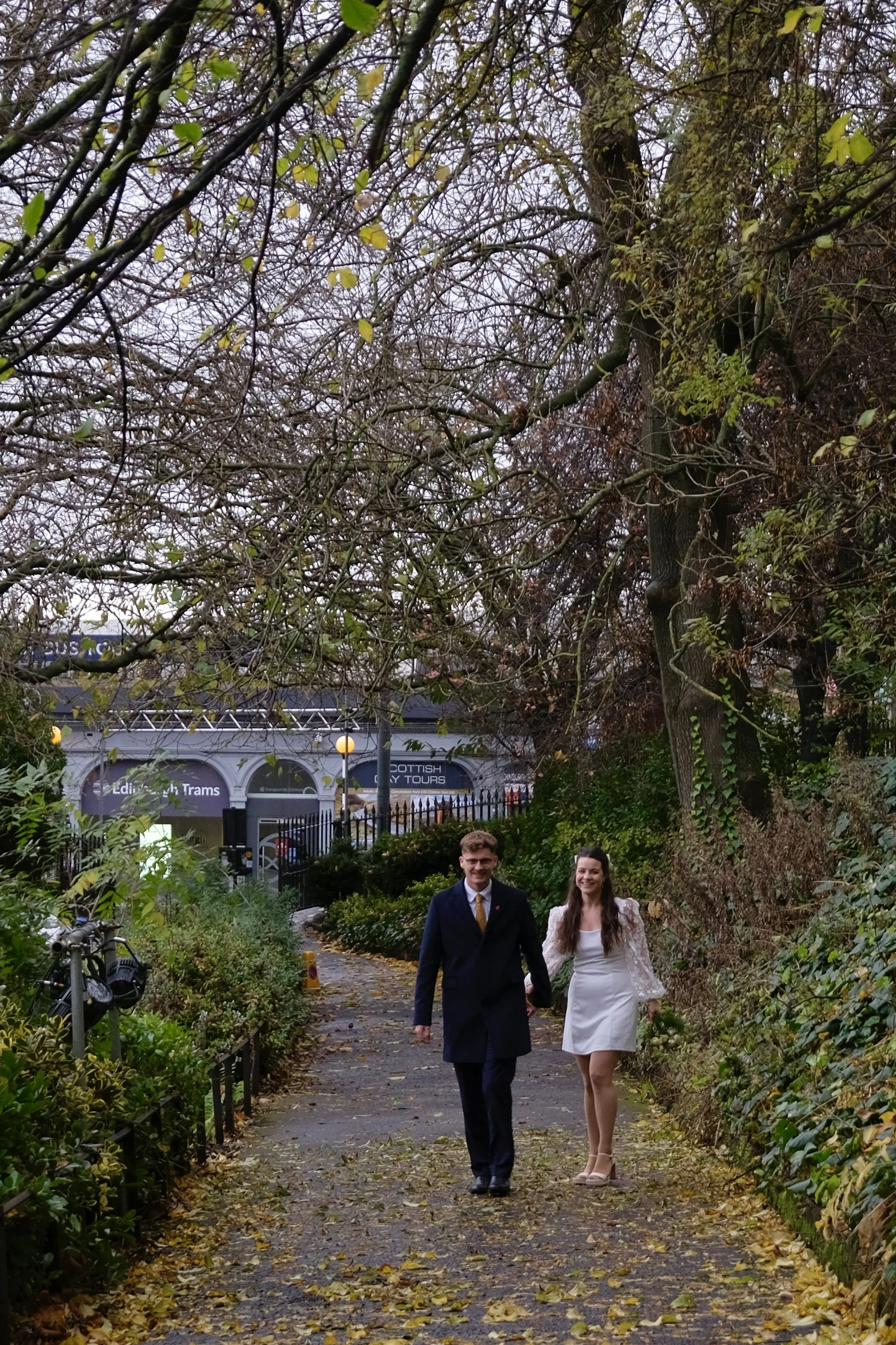 A couple walking hand in hand on a leaf-covered path in a park, trees with sparse foliage, and a sign in the background for Edinburgh Trams.