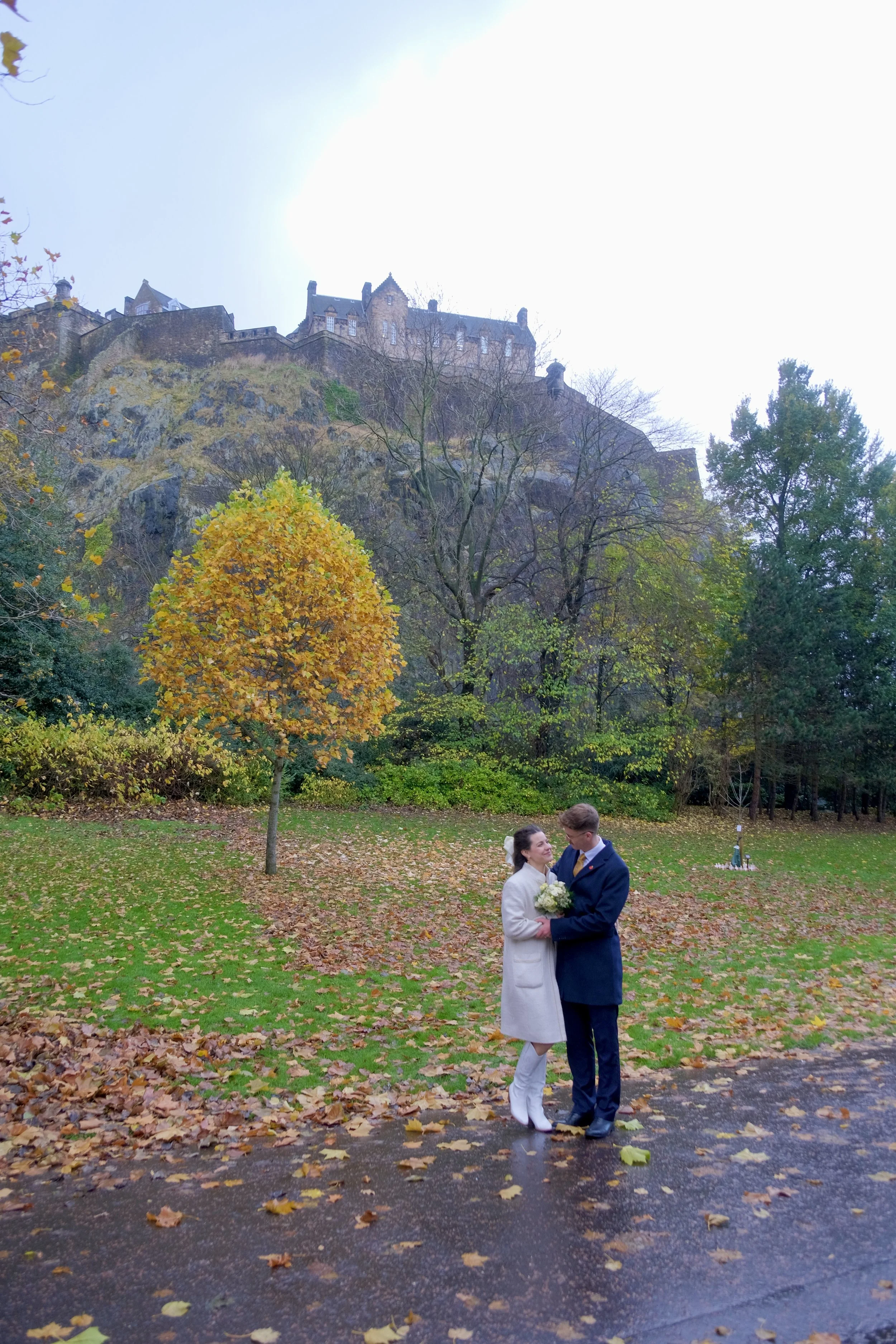 A bride and groom standing together on a sidewalk, holding a bouquet, surrounded by autumn leaves and trees, with a castle on a hill in the background.