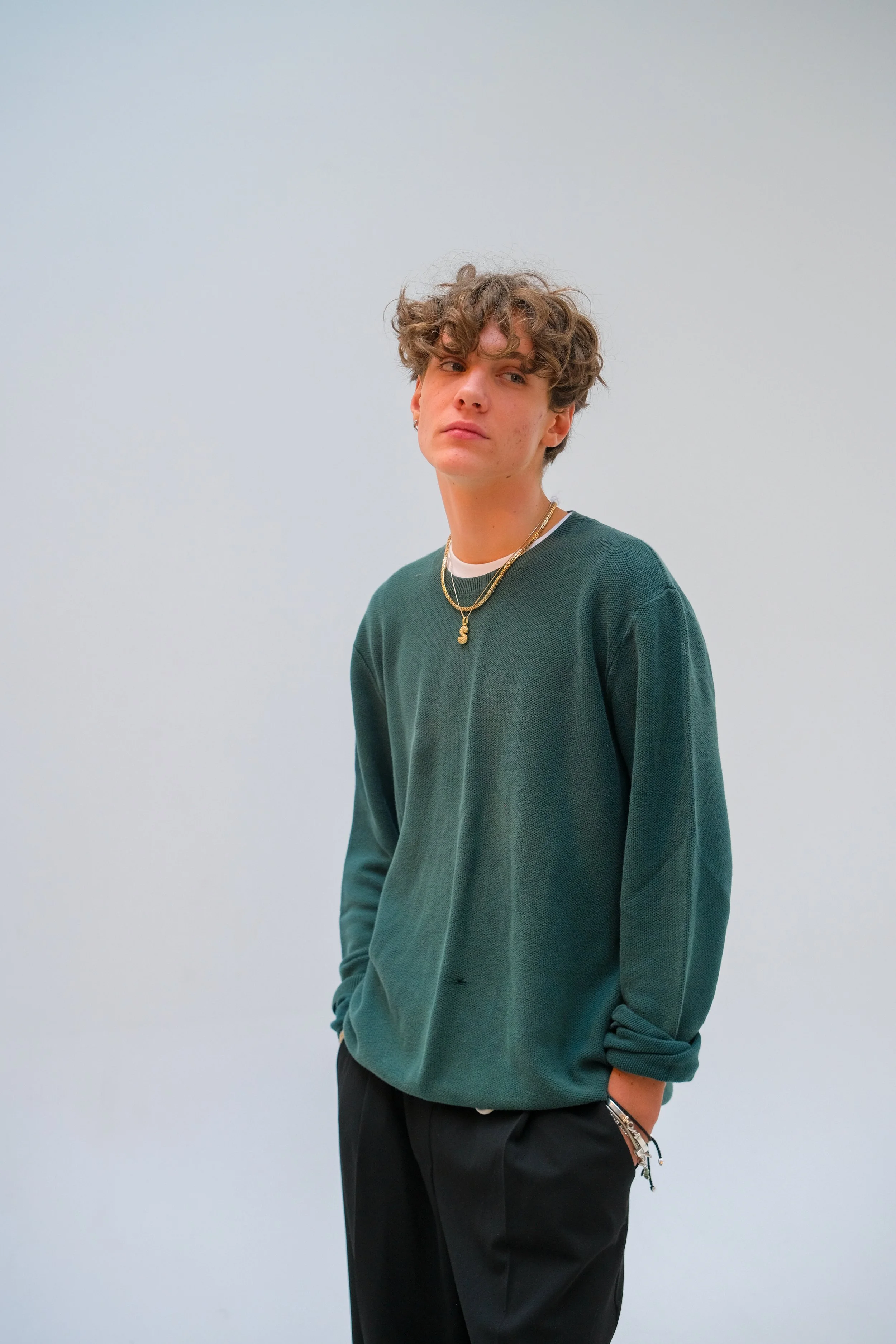 A young man with curly brown hair standing against a plain white background, wearing a dark green sweatshirt, black pants, and layered gold necklaces.