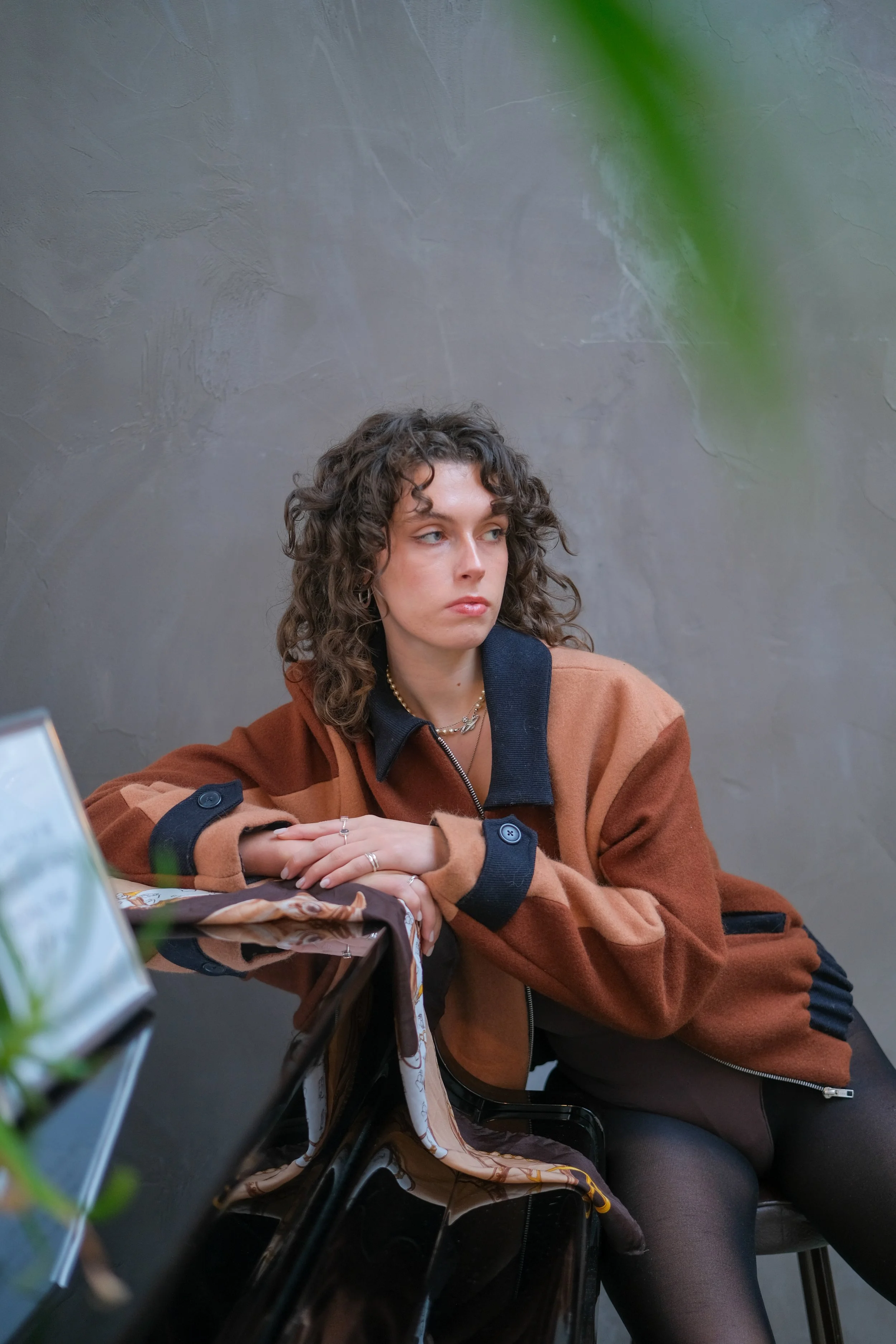 A young woman with curly brown hair wearing a brown and black jacket, sitting at a table with a gray textured wall in the background.