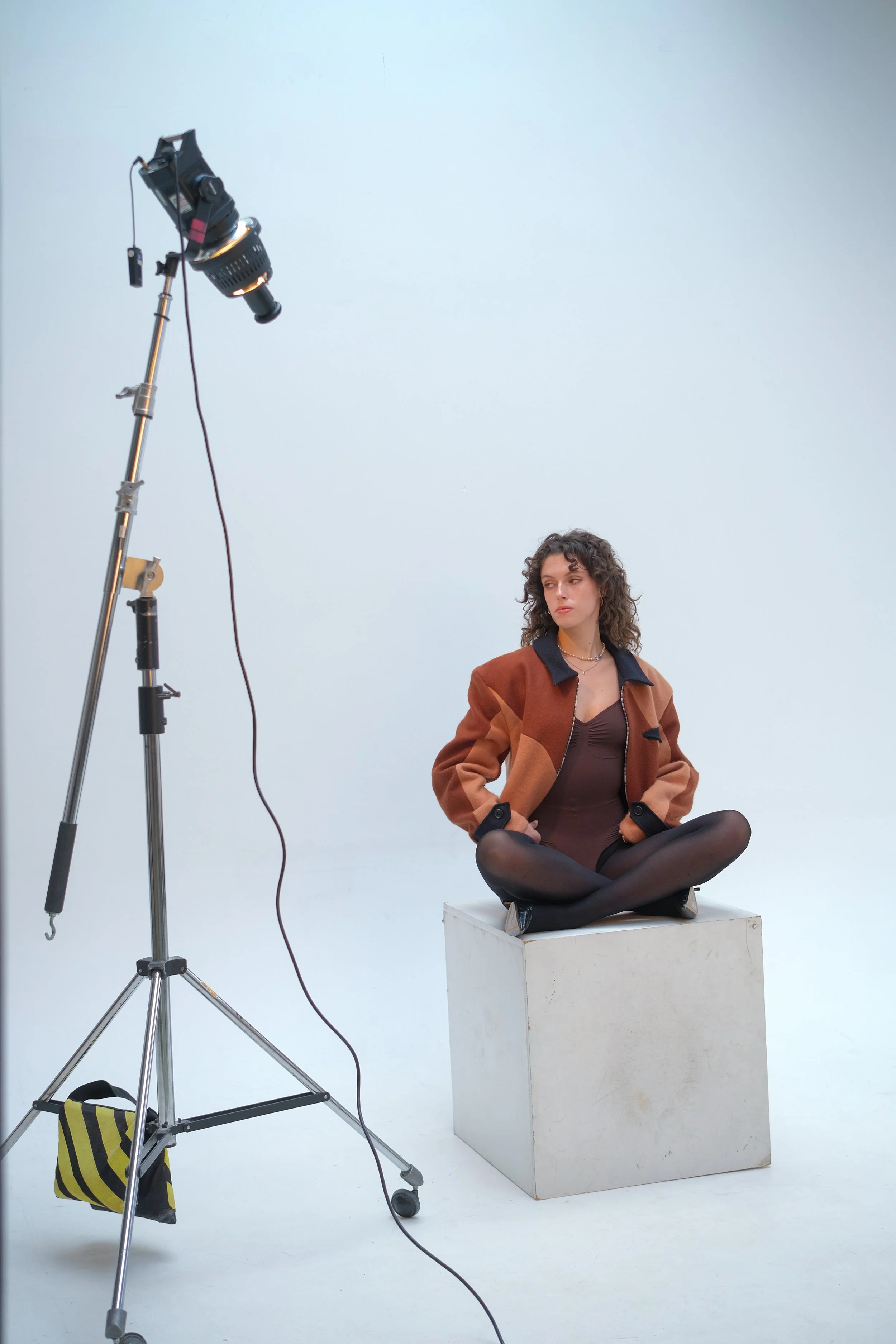 A woman sitting cross-legged on a white block, posed for a photo or video shoot, with professional lighting equipment on a white background.