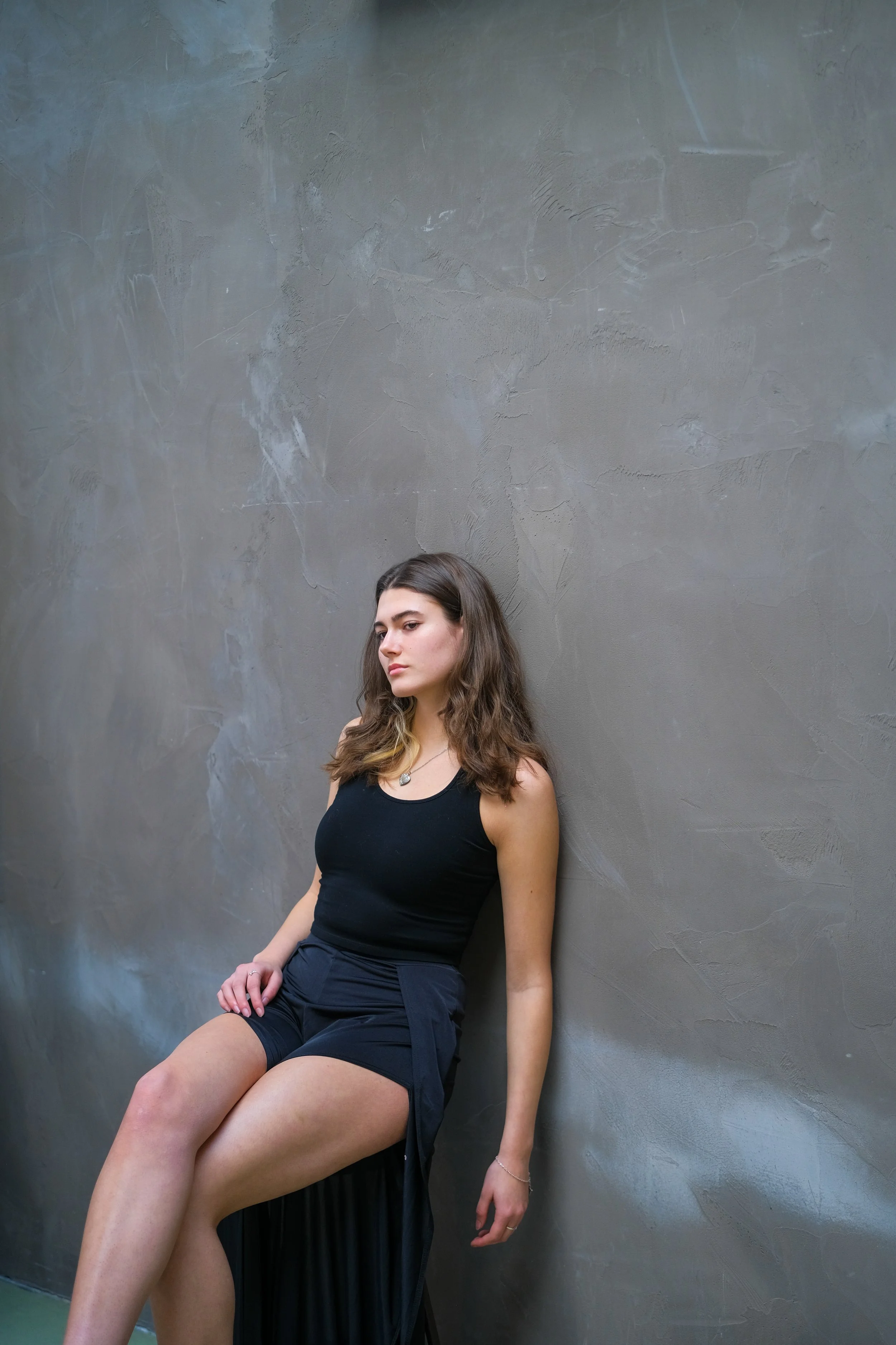A young woman with long brown hair, wearing a black tank top and black shorts, sitting against a textured gray wall with her eyes closed.