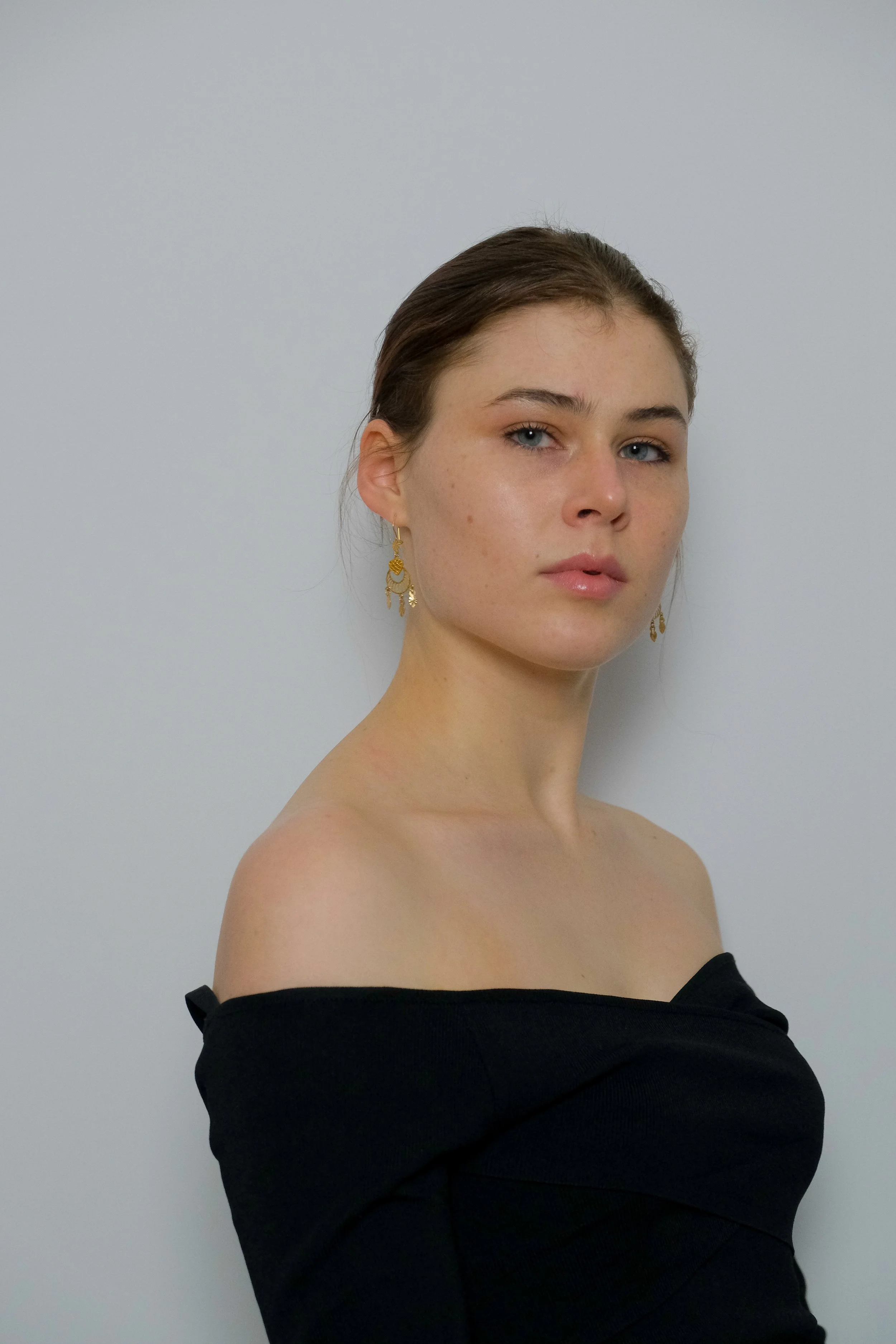 Close-up portrait of a young woman with fair skin, blue eyes, brown hair tied back, wearing gold earrings and a black off-the-shoulder top against a plain white background.