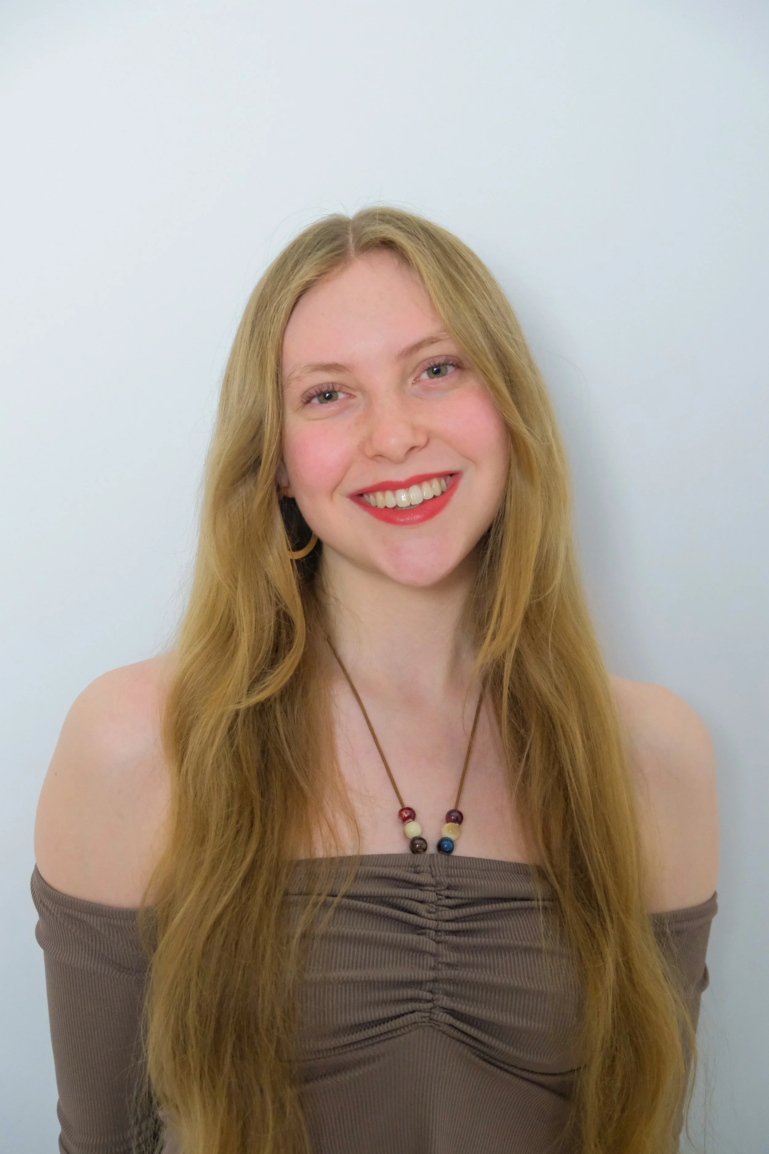 Portrait of a young woman with long red hair, smiling, wearing a brown off-the-shoulder top, a beaded necklace, and hoop earrings, standing against a plain white background.