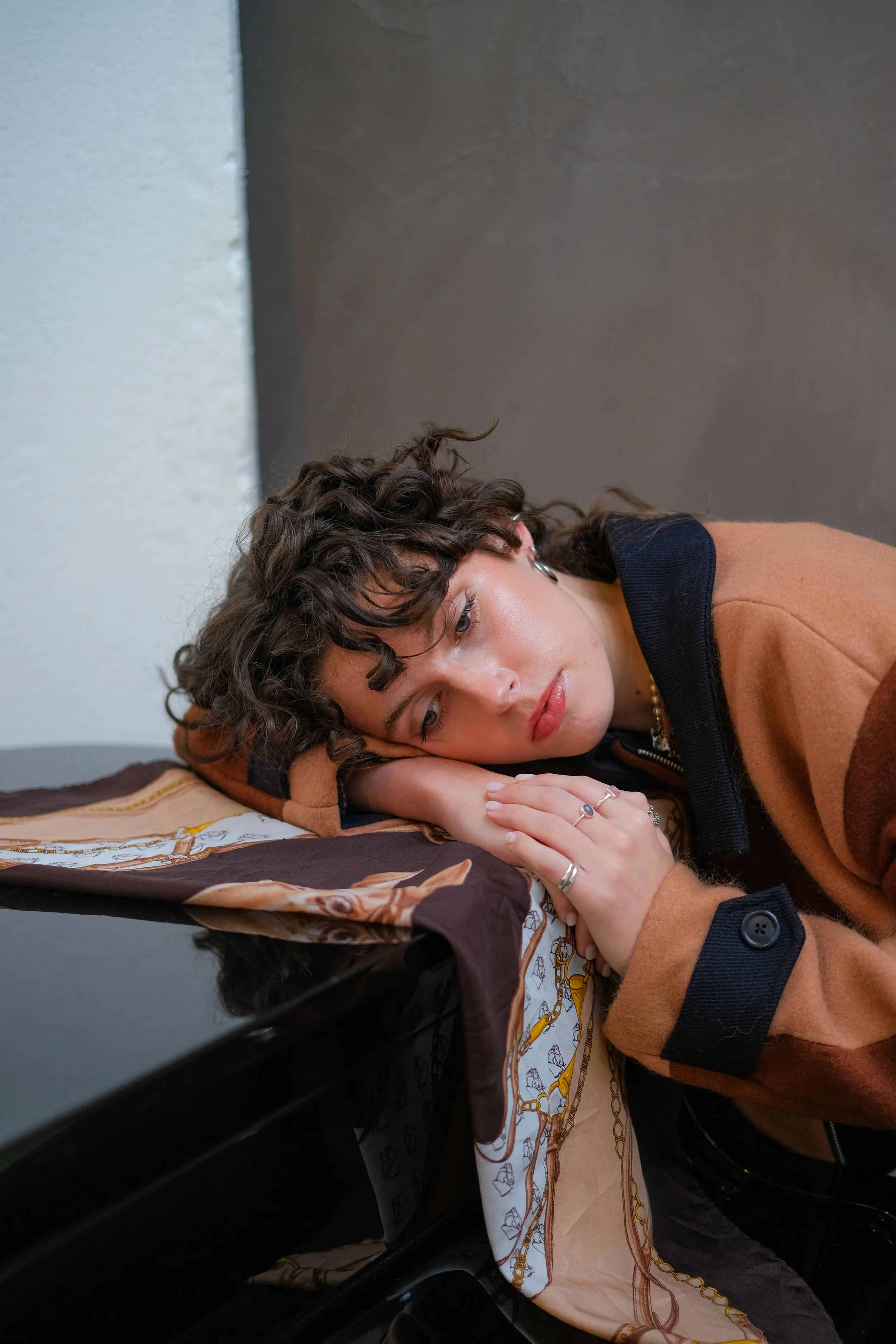 A young woman with curly hair, wearing a brown and navy jacket, is lying on a black table with her head resting on her hands, looking pensively to the side in a neutral-colored room.