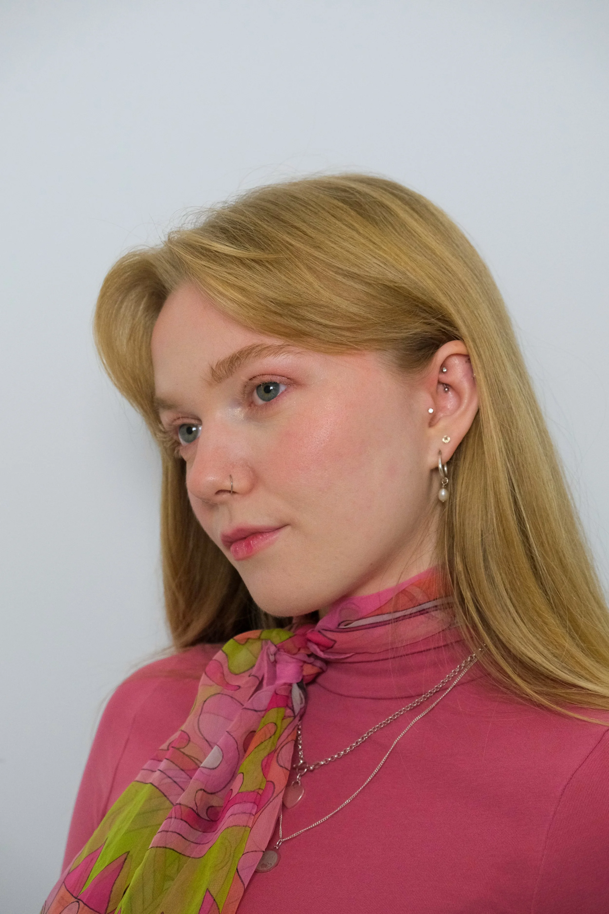 A young woman with blonde hair and blue eyes, wearing earrings, layered necklaces, a pink top, and a colorful scarf tied around her neck, standing against a plain light background.