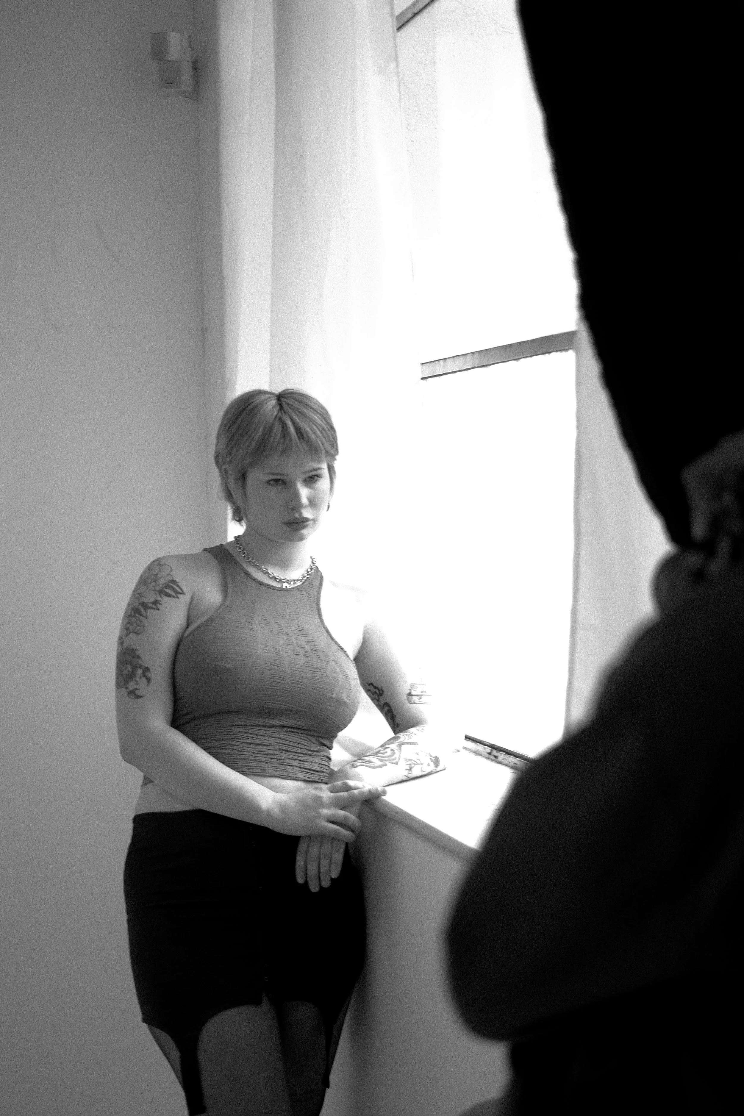 A woman with short hair and tattoos on her arms looks at the camera while standing by a window, with another person partially visible in the foreground.