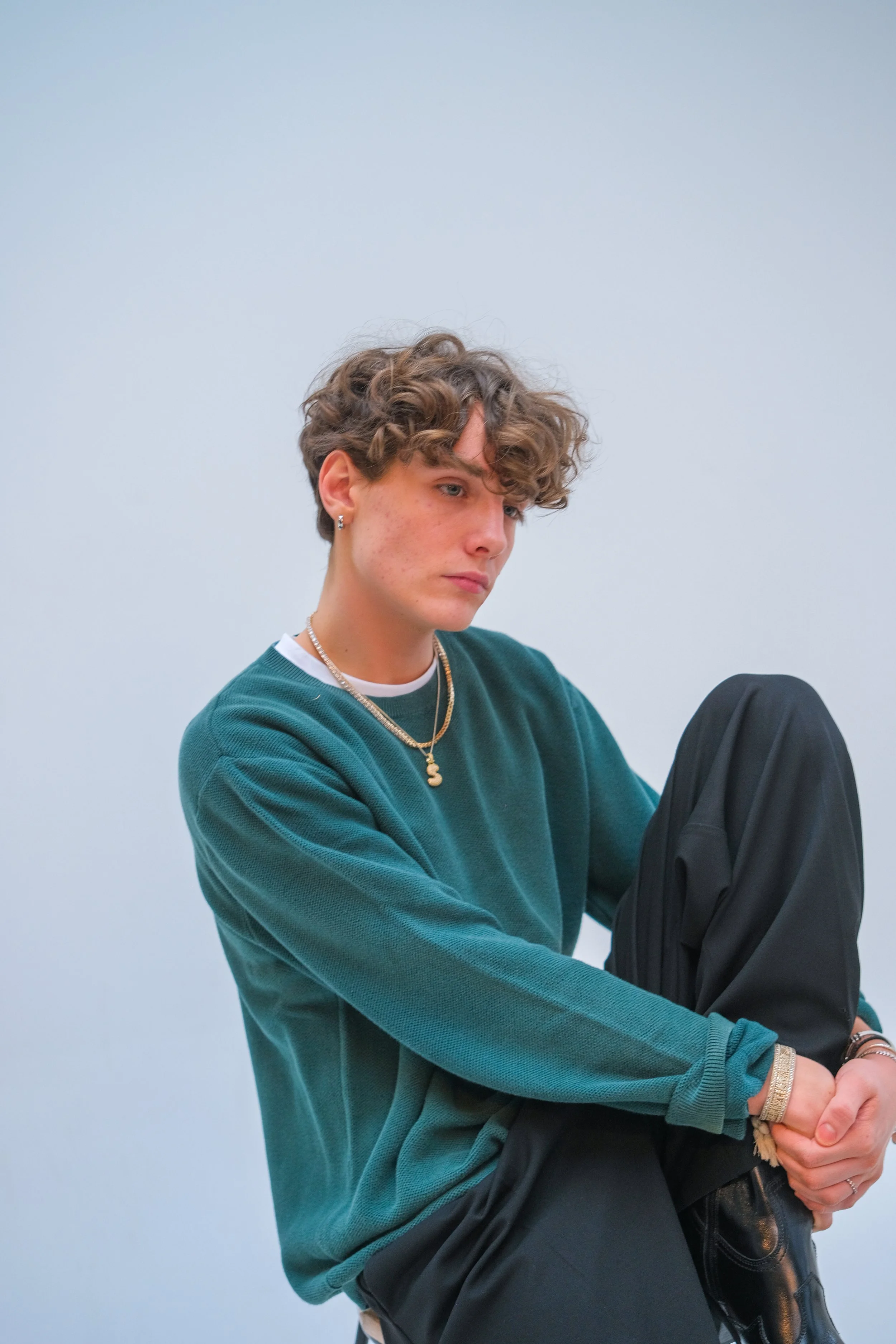 A young person with curly hair, wearing a teal long-sleeve shirt, gold jewelry, and black pants, sitting against a plain light blue background.