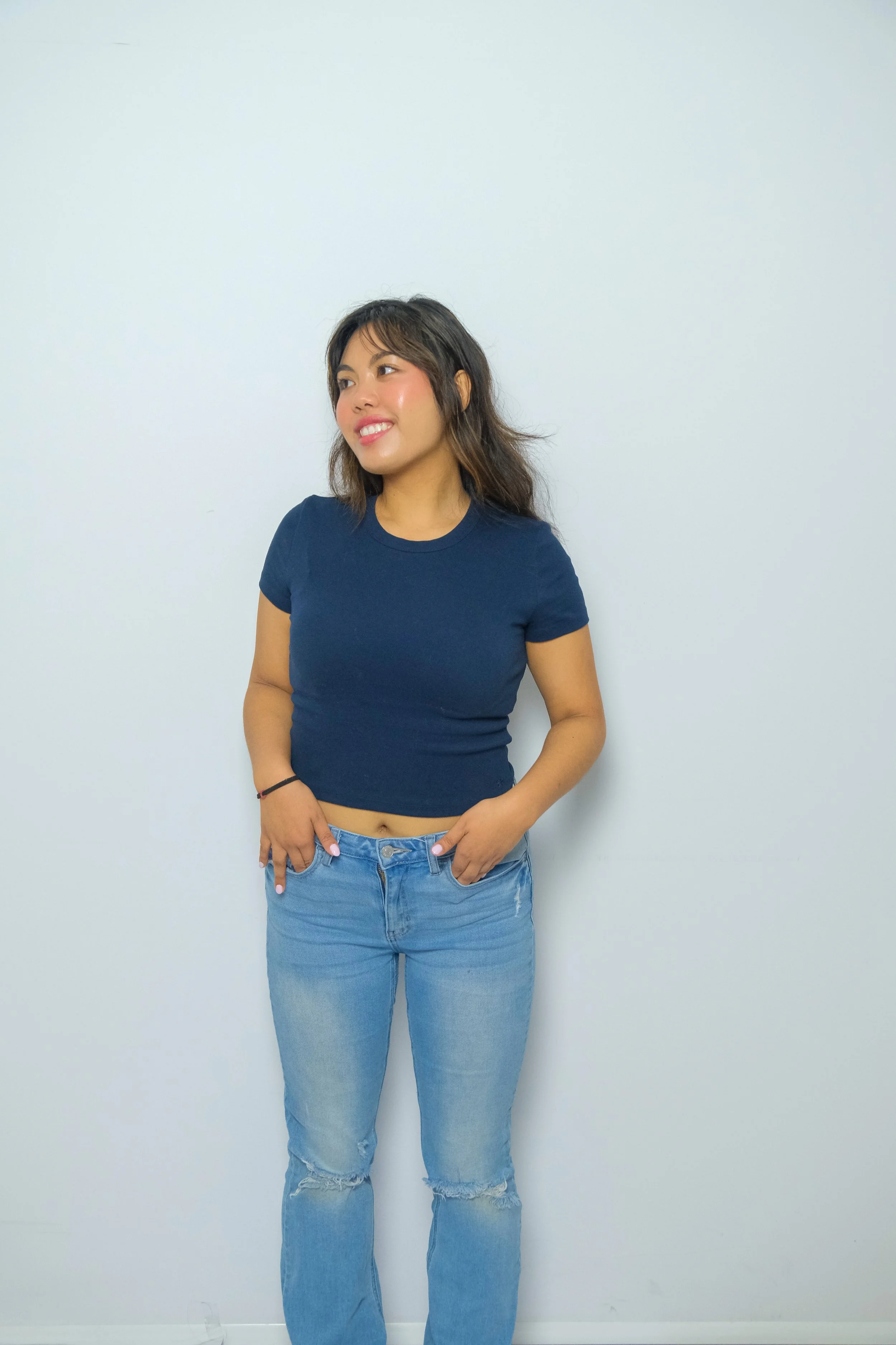 A young woman with wavy hair and a bright smile standing against a plain white wall, wearing a dark blue crop top and light blue ripped jeans.