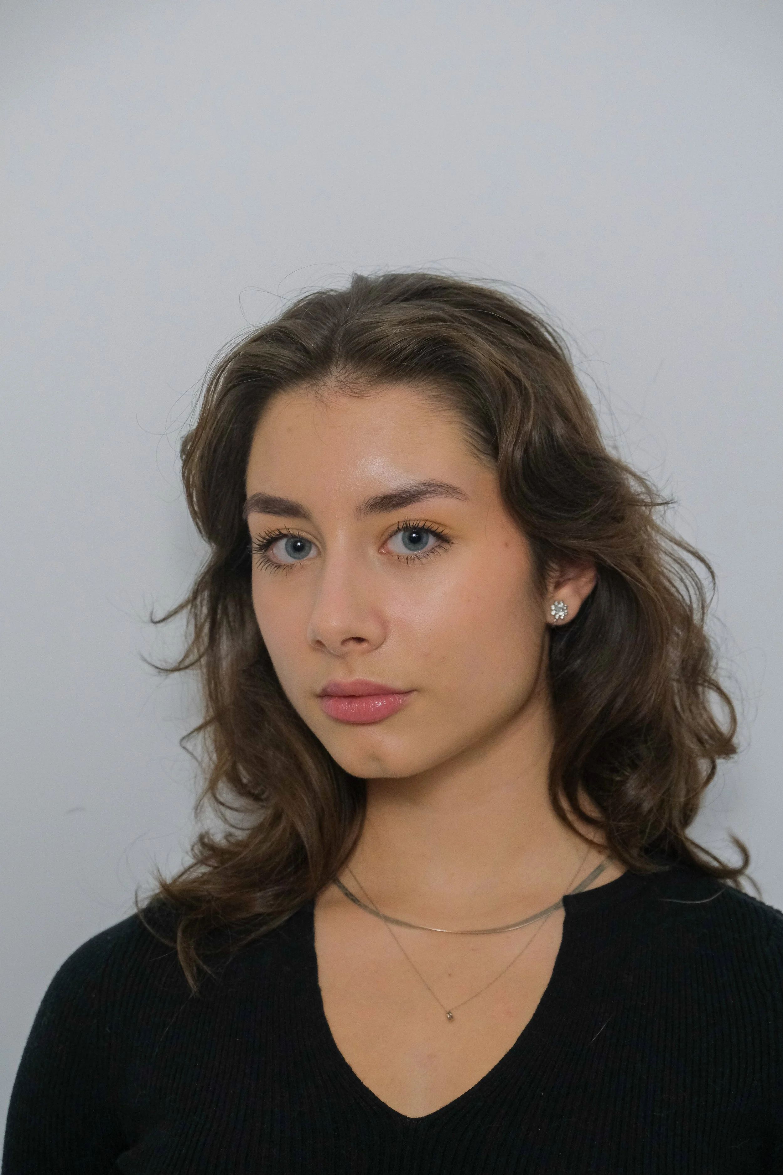 Portrait of a young woman with wavy brown hair, blue eyes, wearing a black top and layered necklaces, against a plain light background.