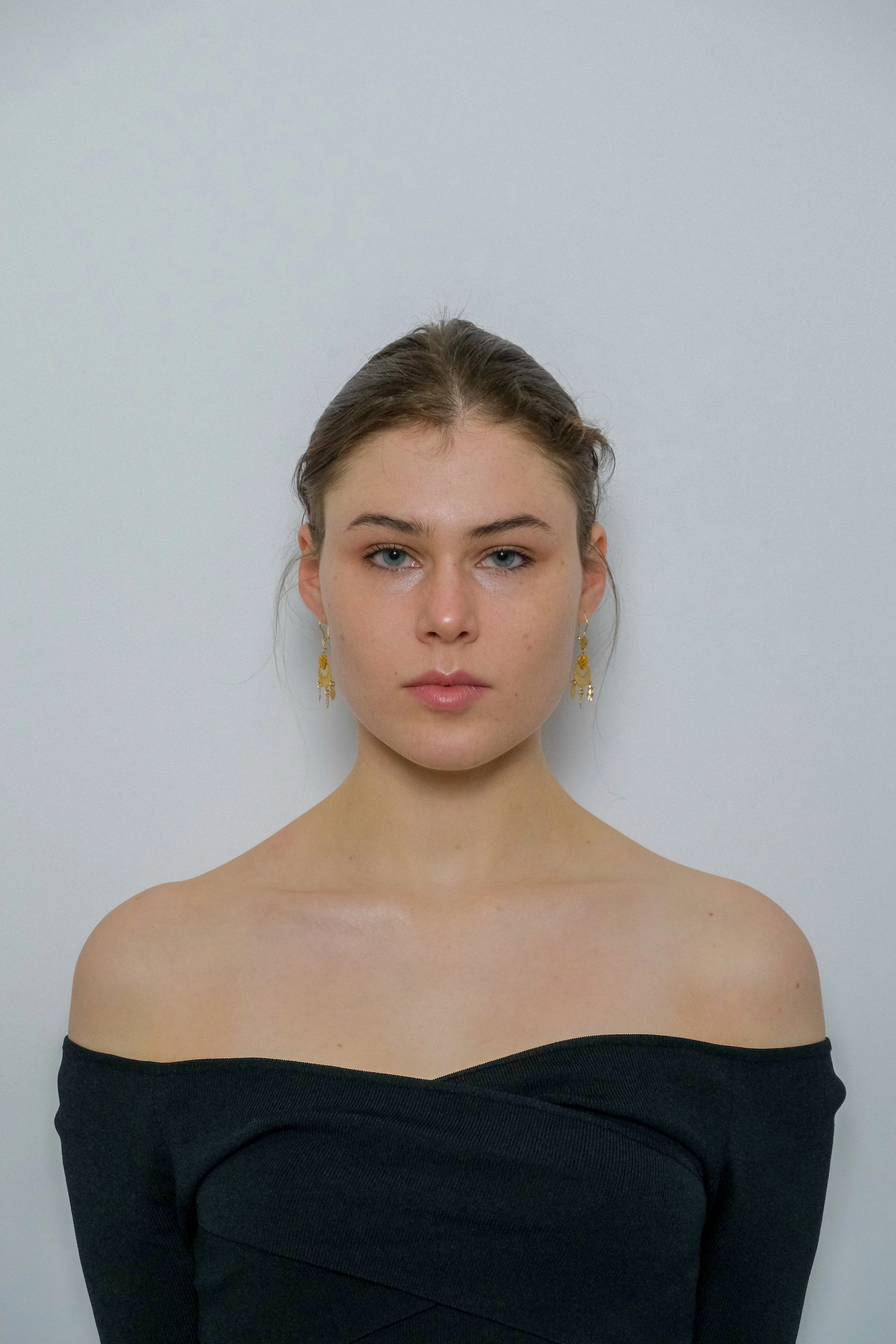 Portrait of a young woman with light skin, blue eyes, and dark hair, wearing gold earrings and a black off-shoulder top, standing against a plain white background.