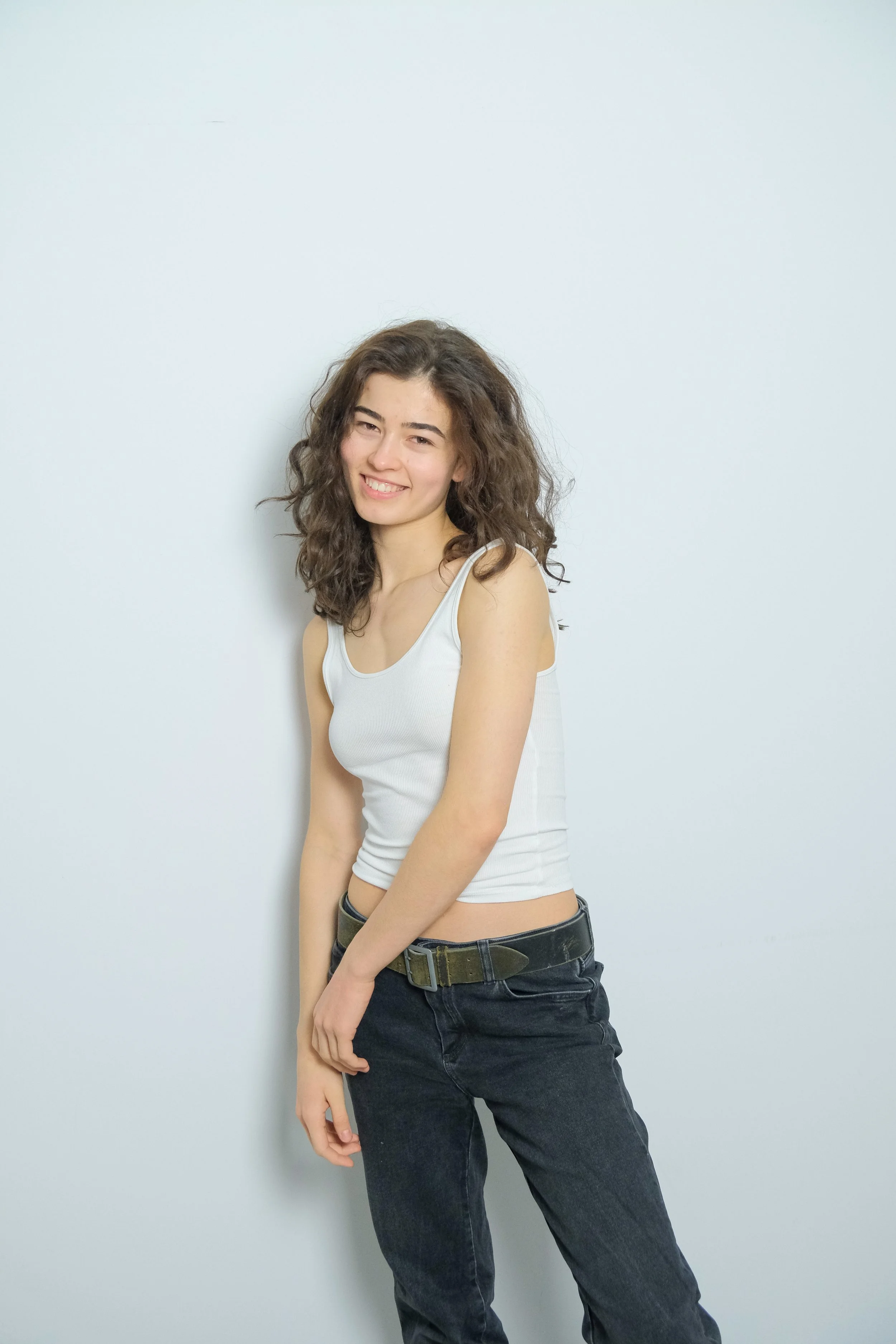 A young woman with curly brown hair smiling and standing against a plain white wall, wearing a white tank top and black jeans.