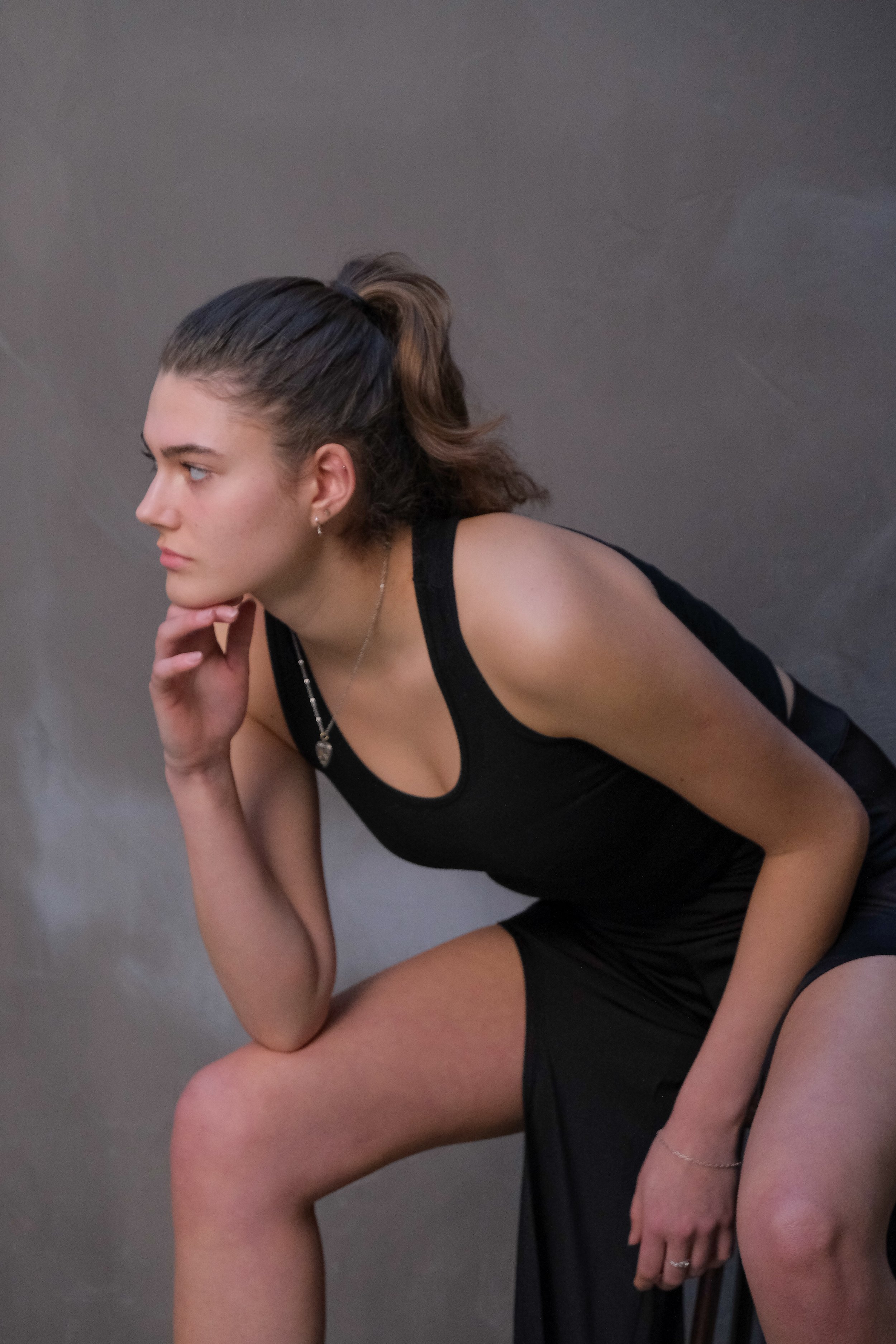 A young woman in black athletic attire is seated in a thoughtful pose, resting her chin on her hand against a plain gray background.