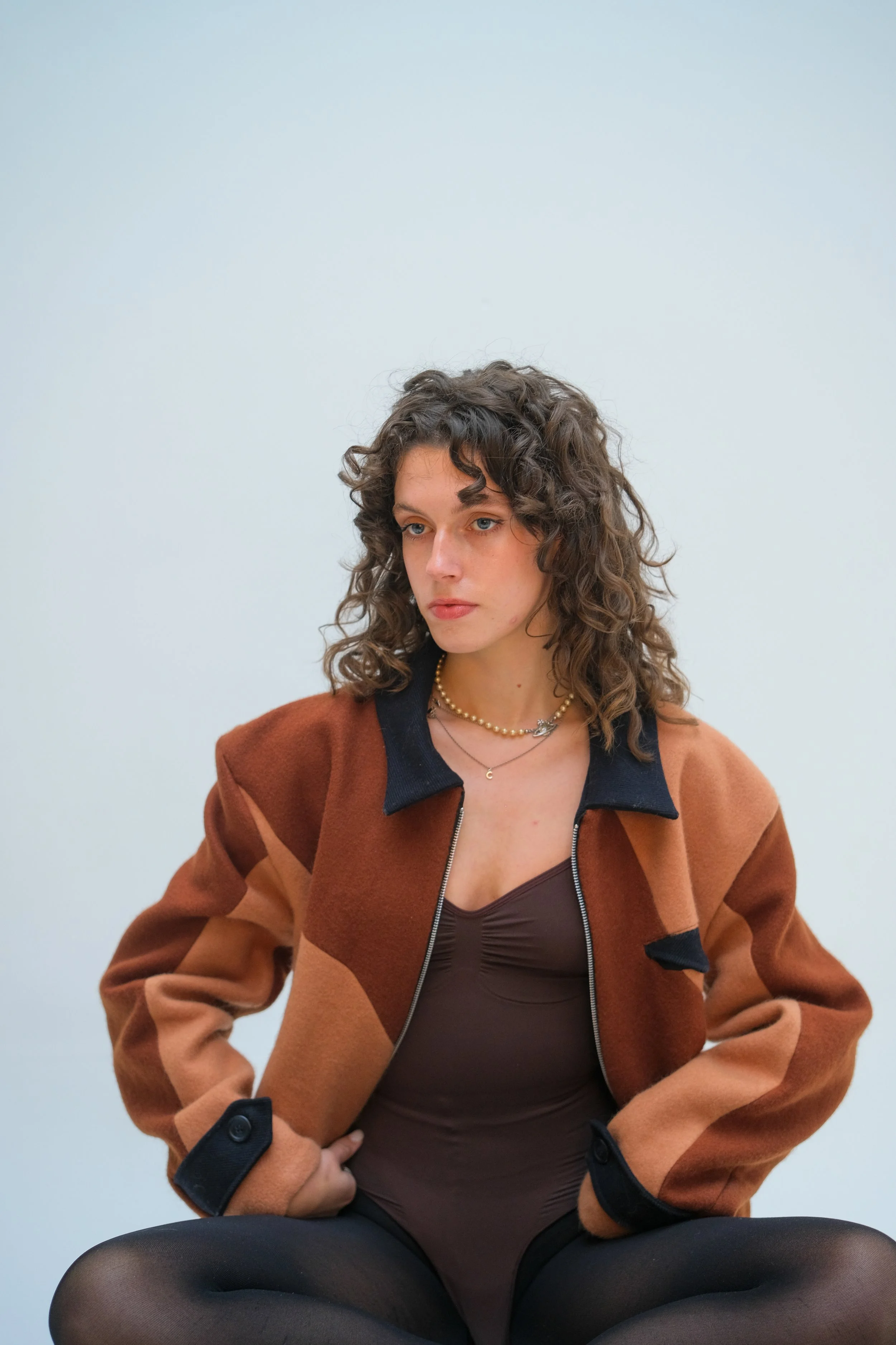 A woman with curly hair wearing a brown jacket over a brown top, sitting with her hands on her hips against a plain light background.