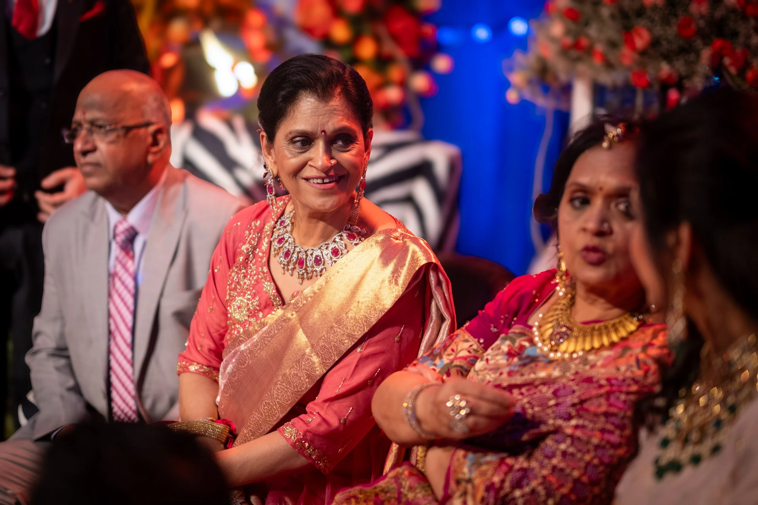 Group of people attending a celebration or wedding, wearing traditional Indian attire, with women in colorful sarees and jewelry, sitting together and engaging in conversation.