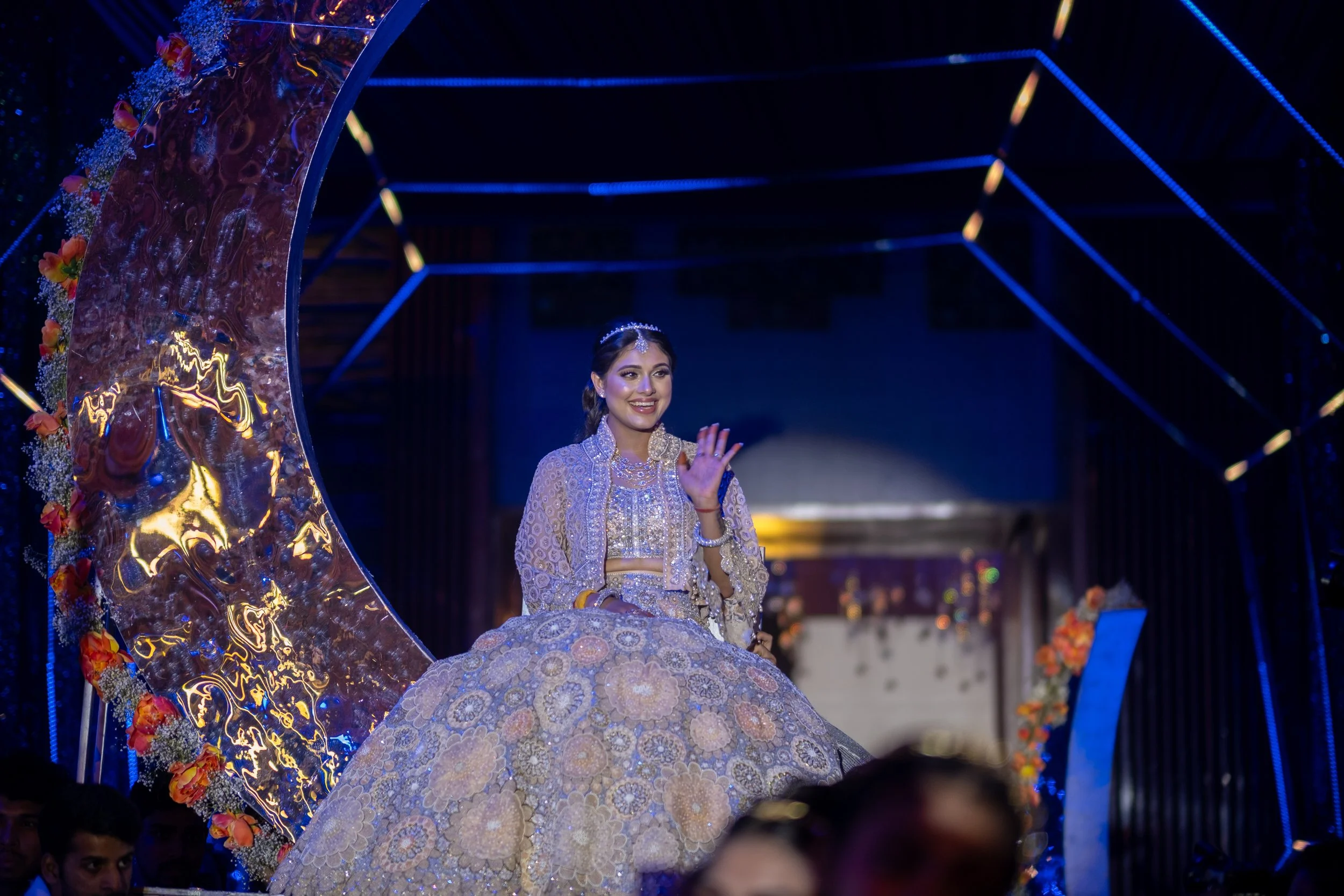 A woman dressed in traditional Indian bridal attire, sitting on a decorated float during a celebration, waving and smiling.