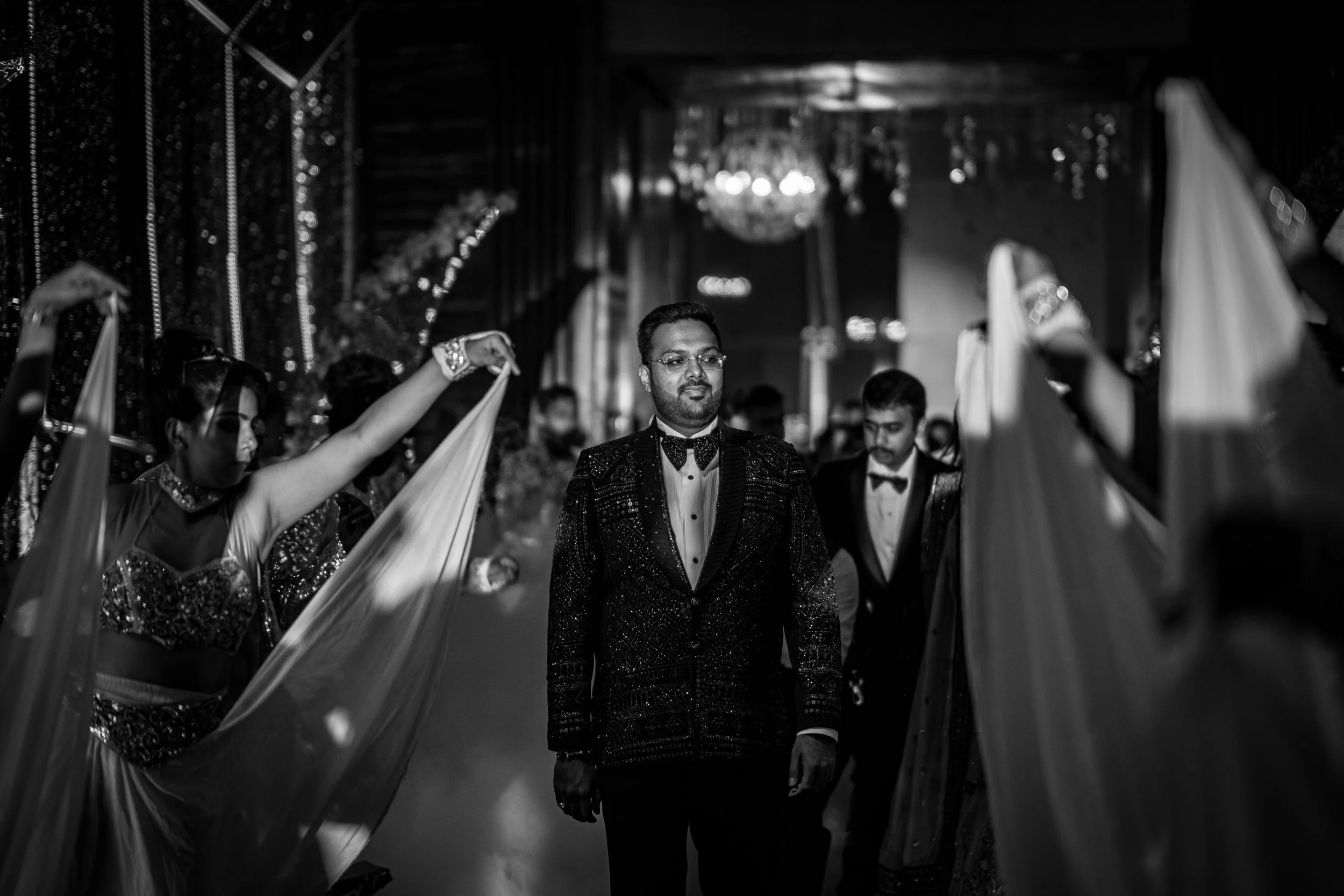 A man in a tuxedo walking through an aisle with women holding fabric drapes on both sides, in a decorated indoor event hall with chandeliers.