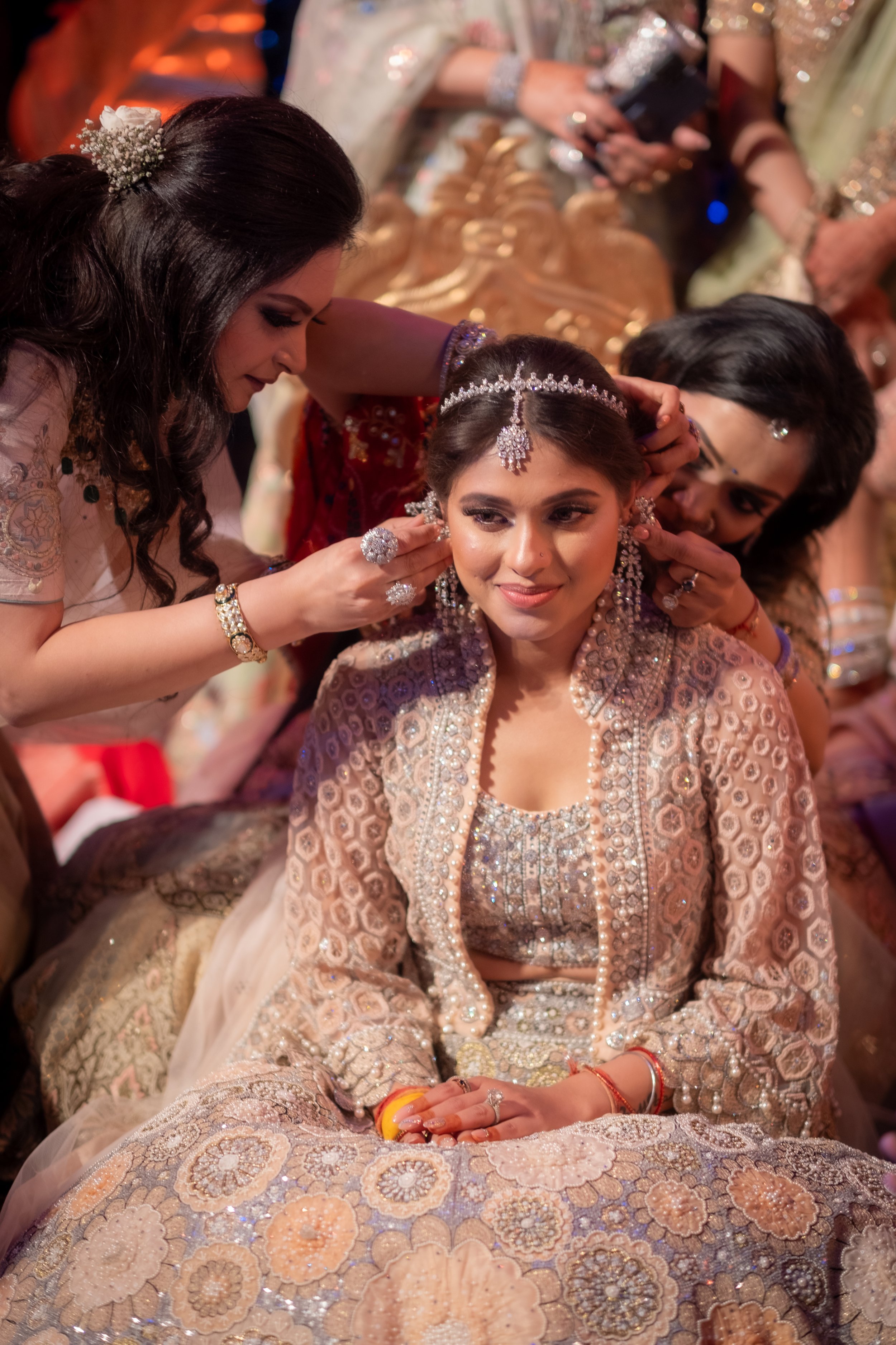 A young woman in traditional Indian bridal attire is being adorned with jewelry by two women on either side of her during a wedding ceremony.