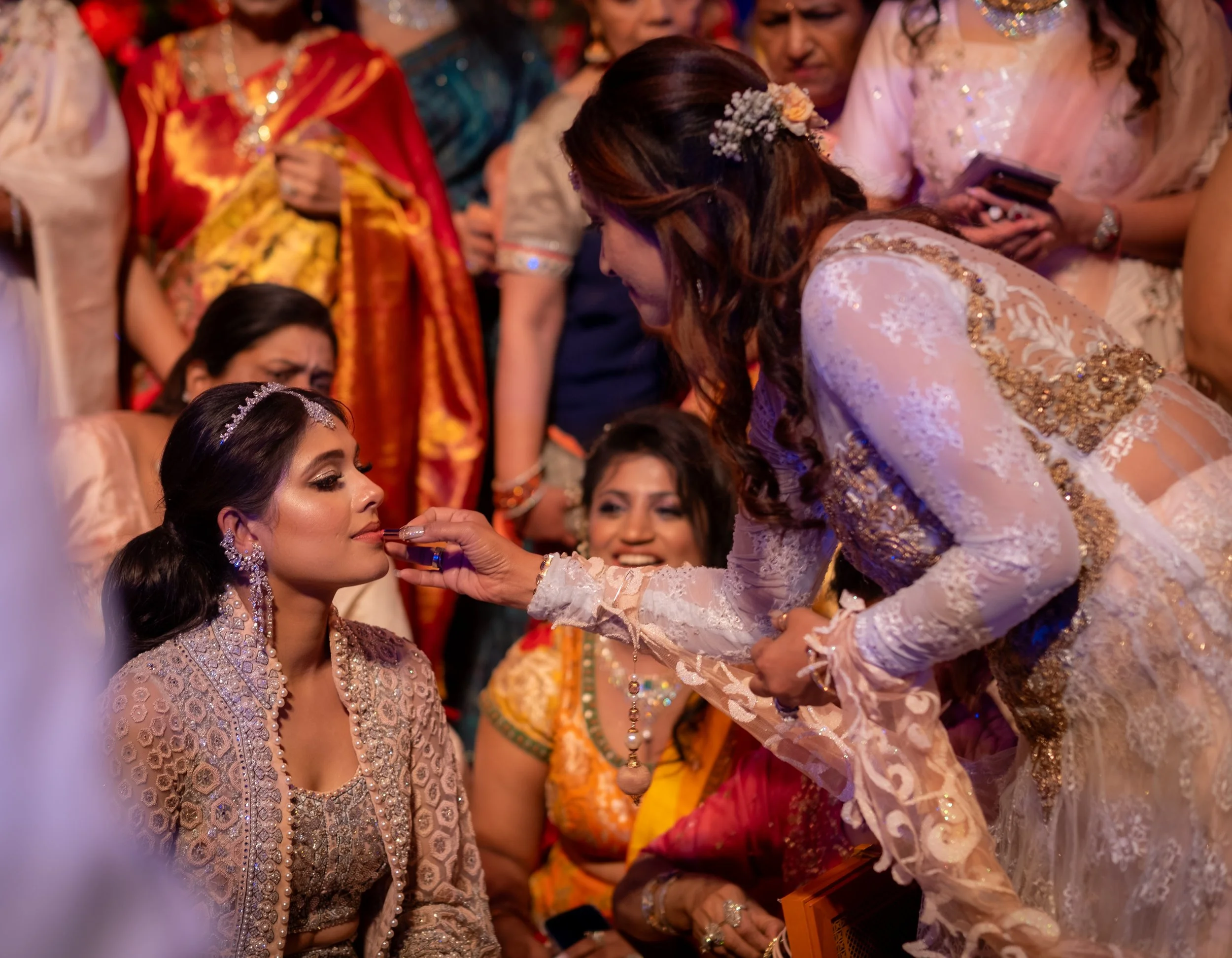 Women dressed in traditional clothing participating in a cultural ceremony, with one woman applying lipstick or makeup to another woman in a group setting.