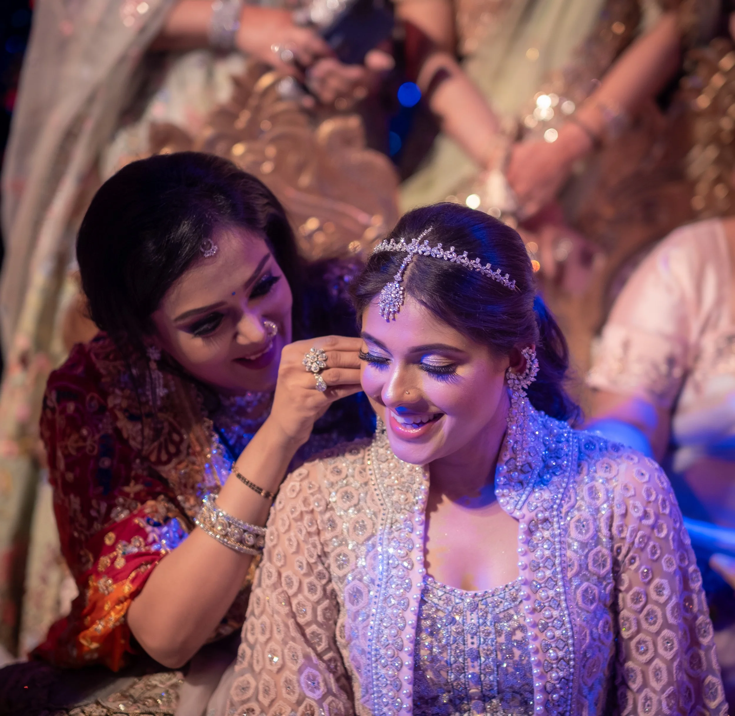 A bride and a woman, presumably her relative or friend, are getting ready during a traditional Indian wedding. The bride is smiling with her eyes closed, wearing ornate jewelry and a heavily embellished outfit, while the other woman is adjusting her 
