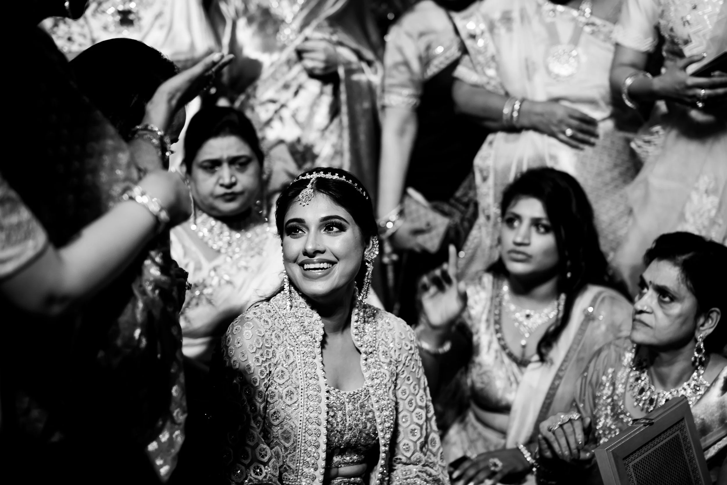Black and white photo of a joyful woman surrounded by women dressed in traditional Indian attire, with jewelry and accessories, during a celebration.