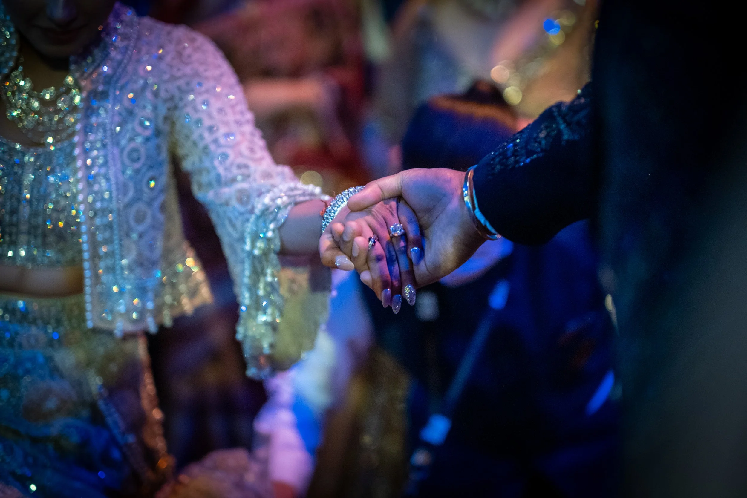 A close-up of two people holding hands at a celebration or party, with one person wearing a detailed, sparkly outfit and jewelry, and the other in a dark textured outfit.