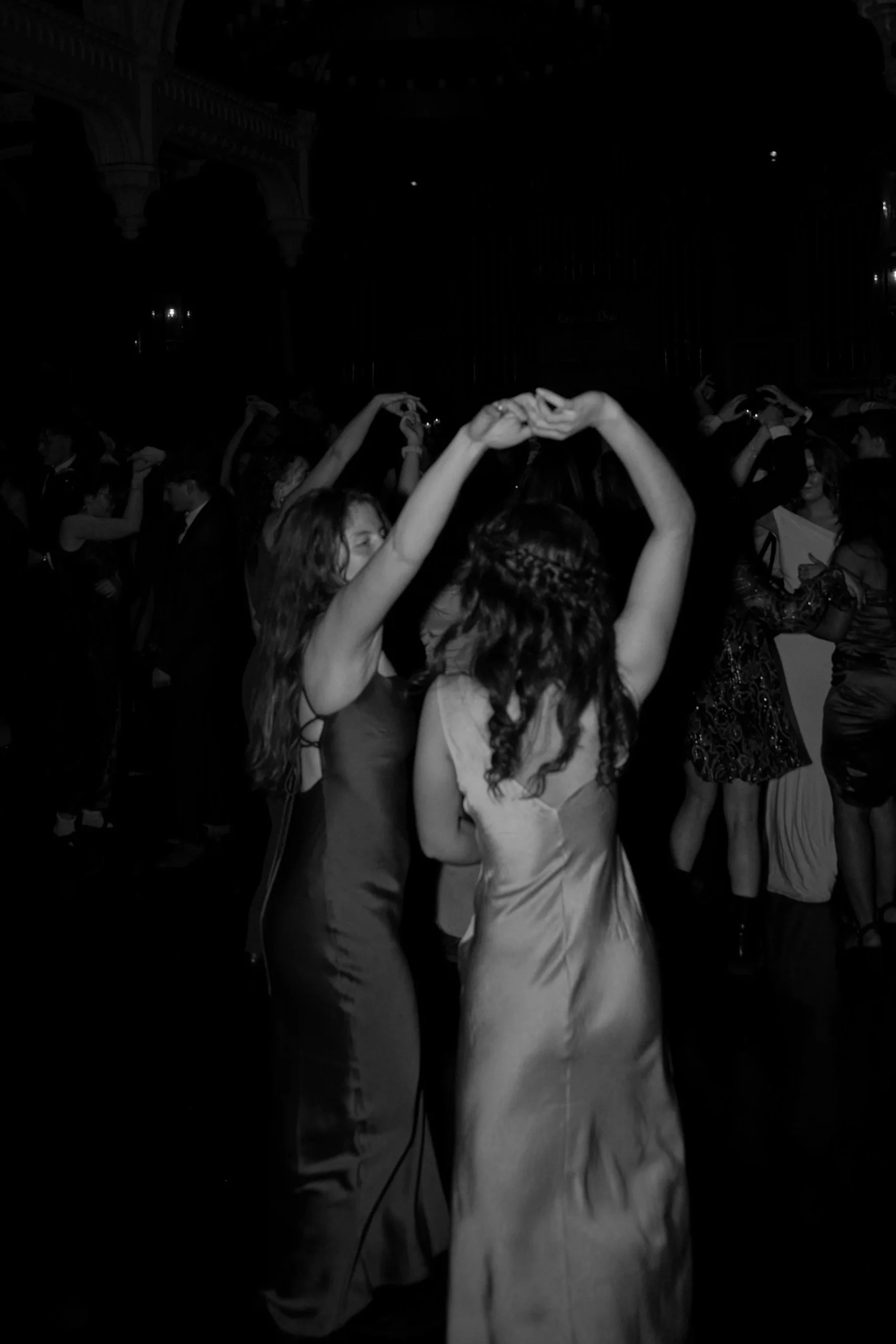 Two women dancing closely at a formal event or dance party, holding hands above their heads, with other people dancing in the background.