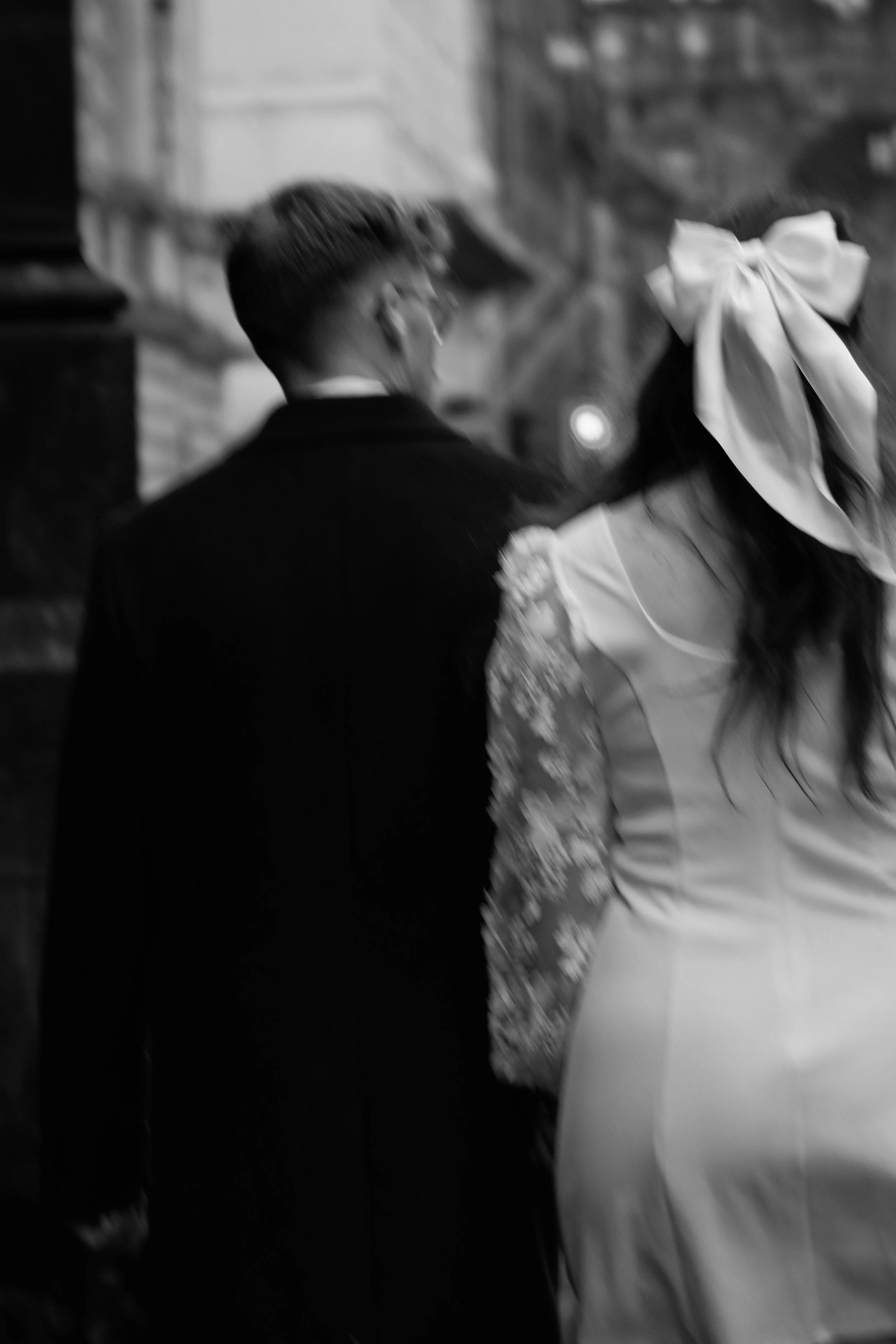 Black and white photo of a couple in formal attire, walking away from the camera inside a building with stone walls. The woman has long hair and a large bow in her hair; the man is wearing a dark suit.