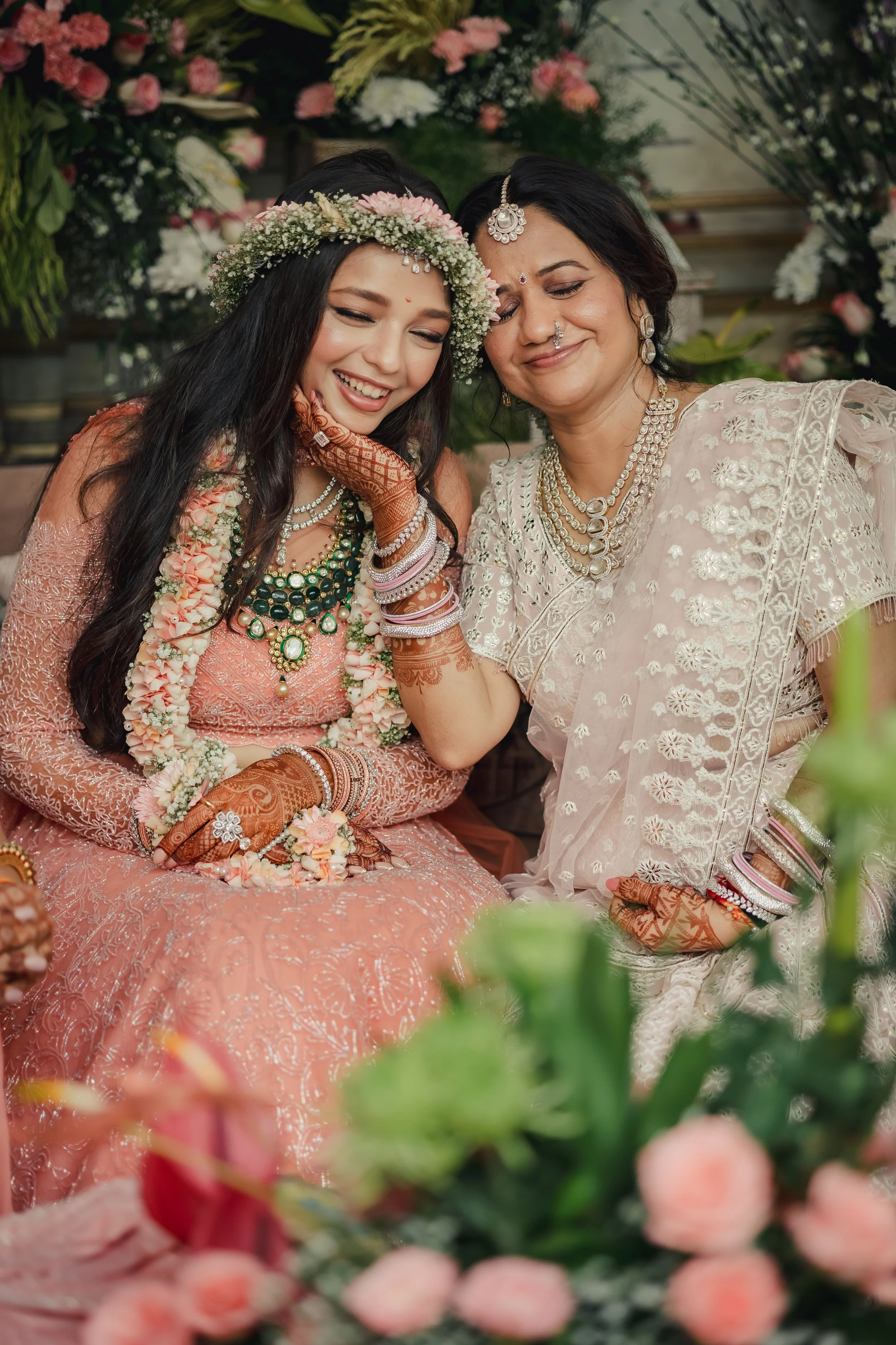 A bride in a pink traditional dress and a woman in a white saree smiling and sharing a happy moment at a wedding decorated with flowers.