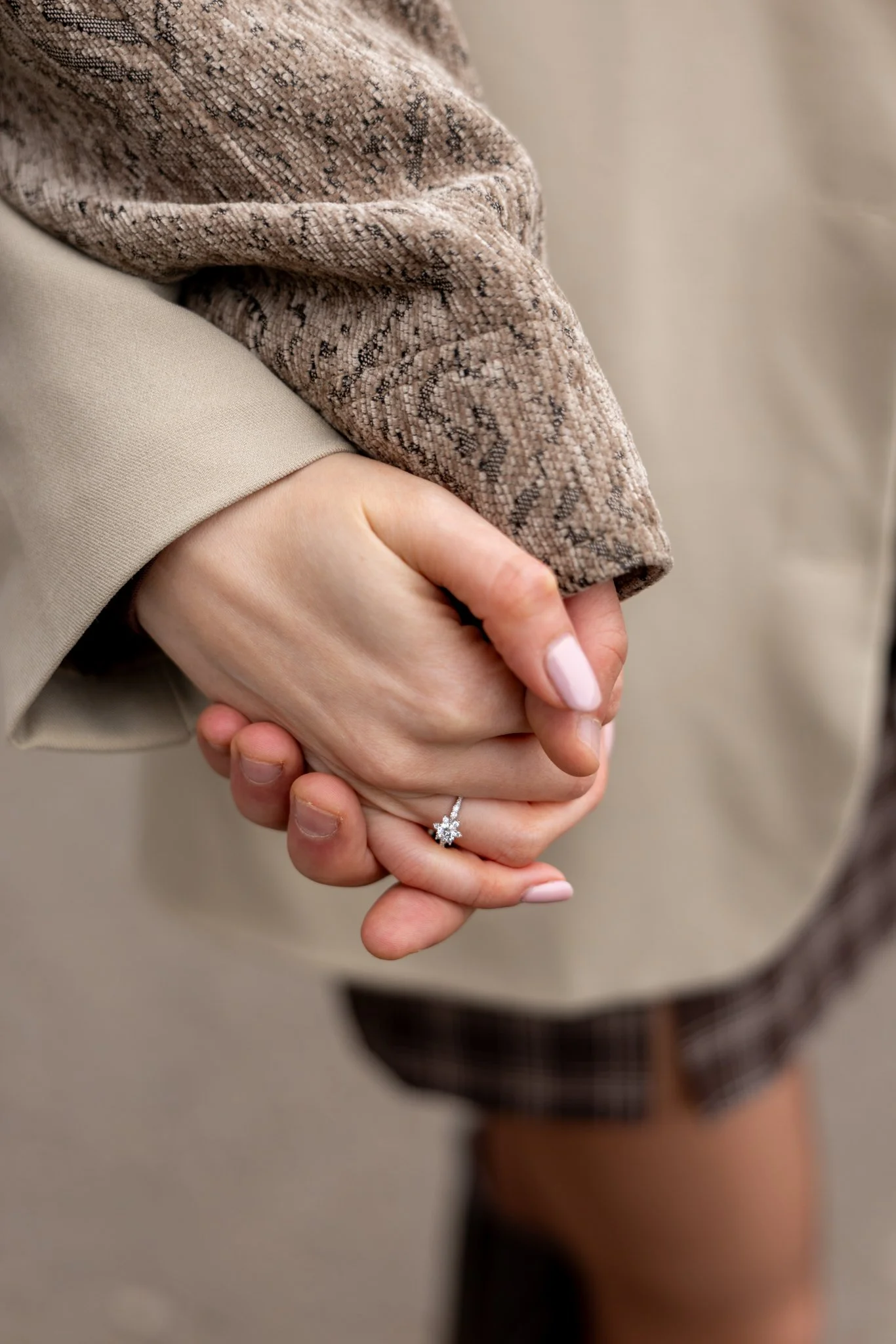 Close-up of two people holding hands, one wearing a wedding ring with a star-shaped diamond, the other with light pink painted nails. They are wearing beige and woven patterned jackets.